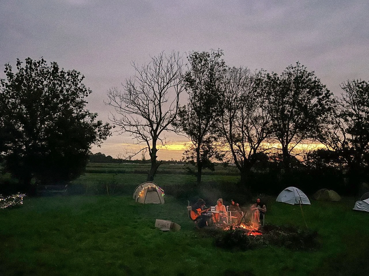 Eine Gruppe von Menschen sitzt um ein Lagerfeuer auf einem Feld, umgeben von Bäumen und Zelten. Es ist Dämmerung, und der Himmel ist bewölkt.