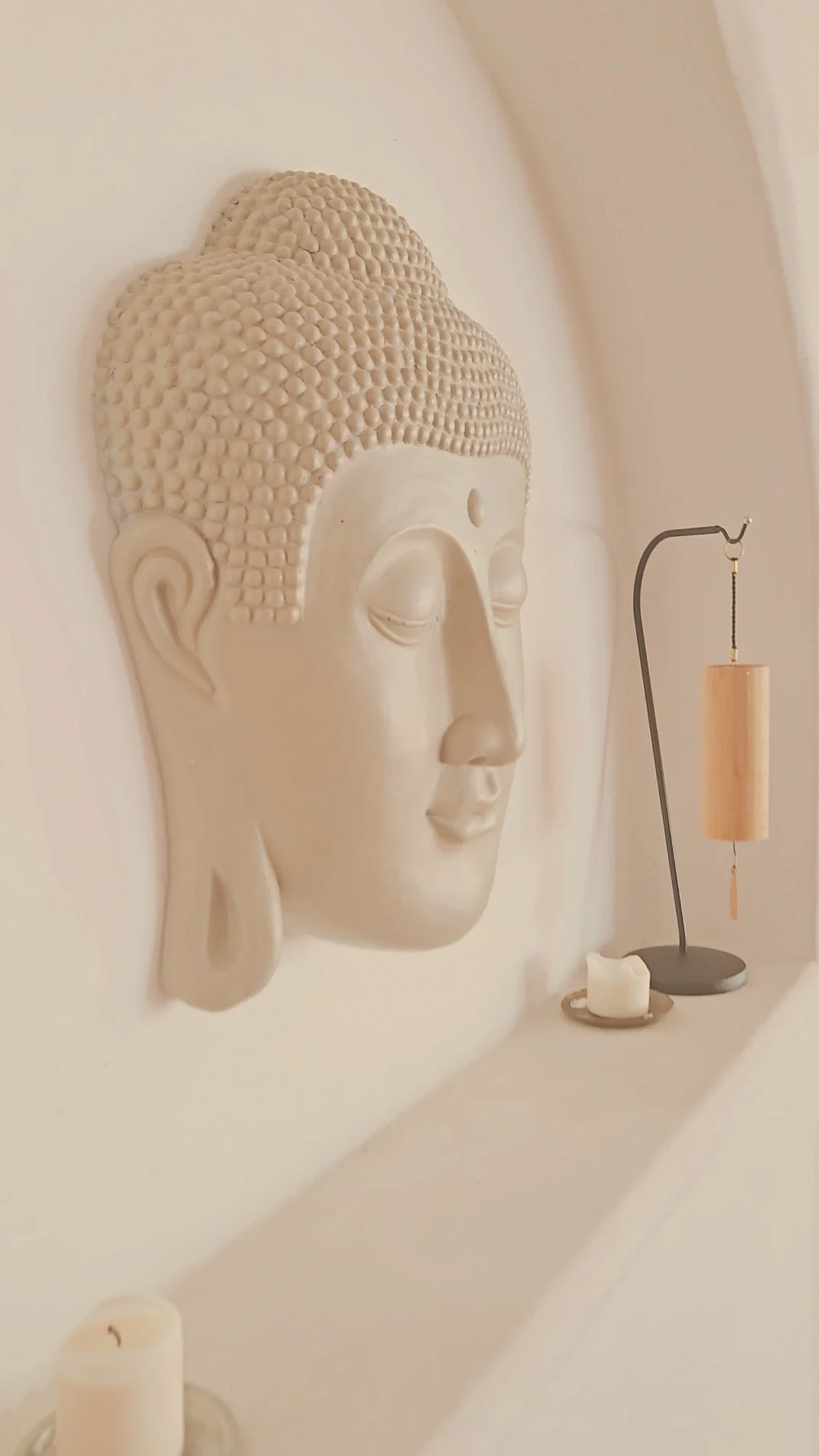 Weiße Buddha-Kopf-Skulptur an der Wand neben einer dekorativen Kerze und einem Holz-Windspiel.