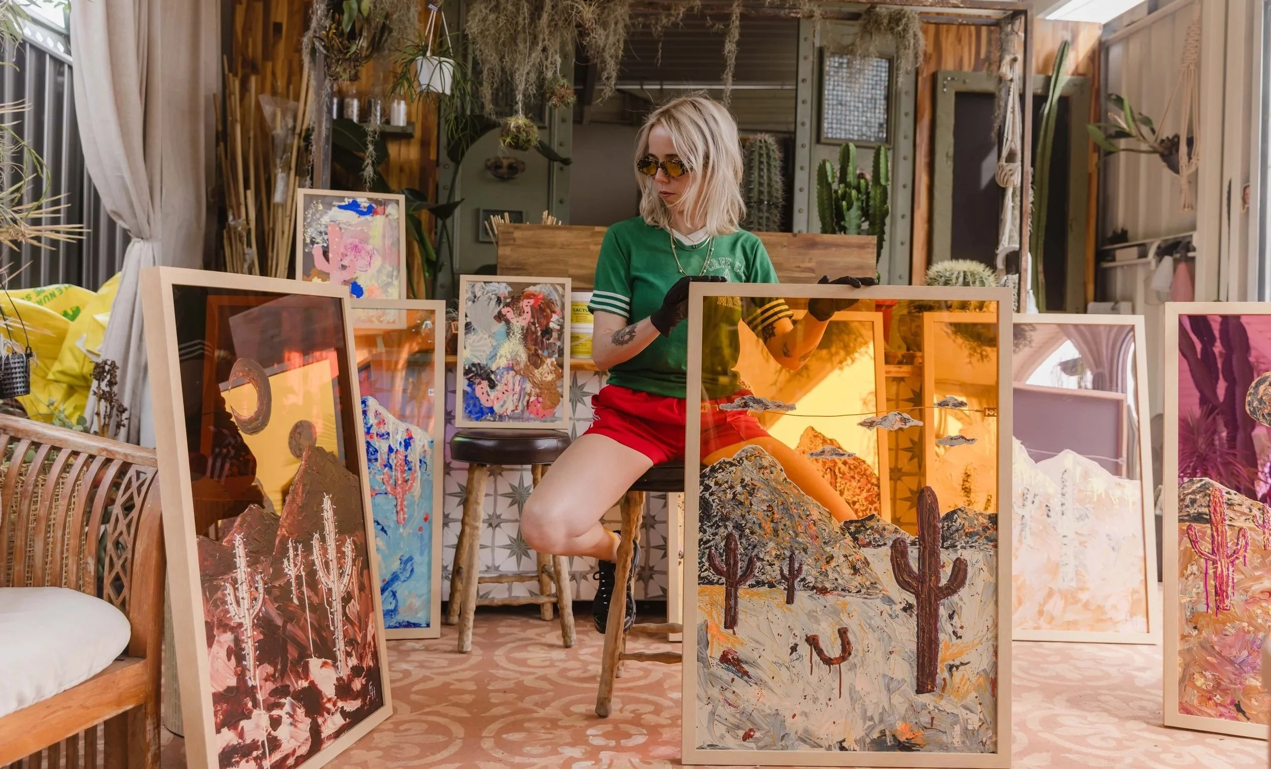 A woman with blonde hair, wearing yellow-tinted sunglasses, a green t-shirt, red shorts, and black gloves, sitting on a wooden stool inside an art studio or gallery. She is surrounded by colorful paintings of desert scenes with cacti and mountains, s