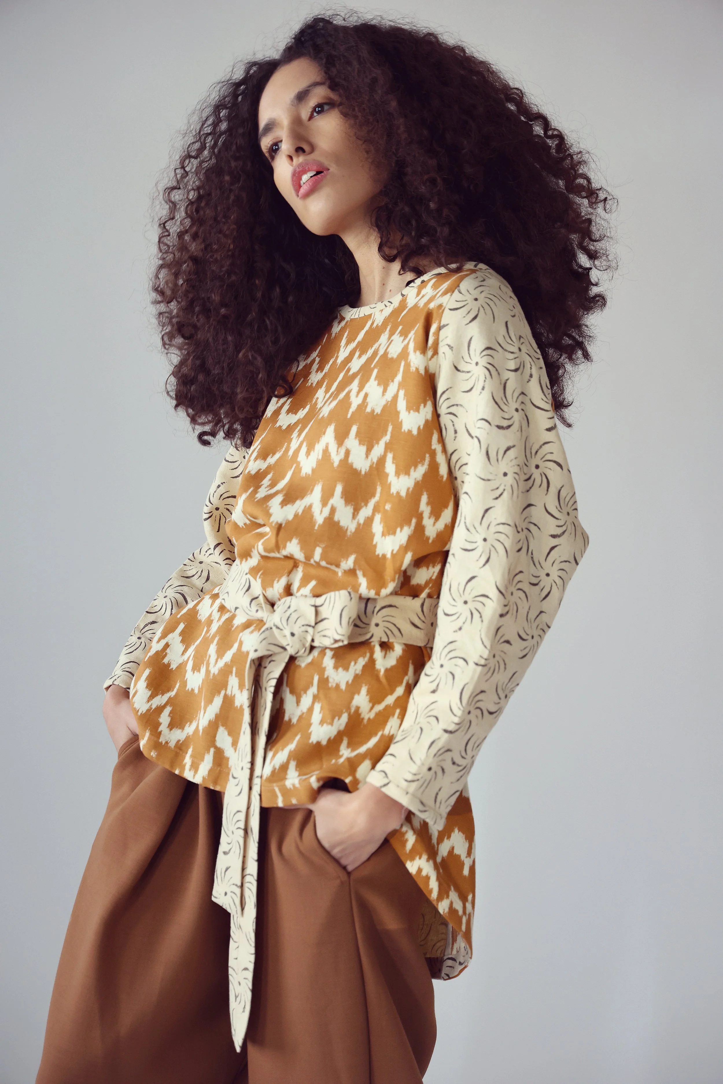 A woman with curly hair wearing a patterned beige and tan top and loose brown pants.