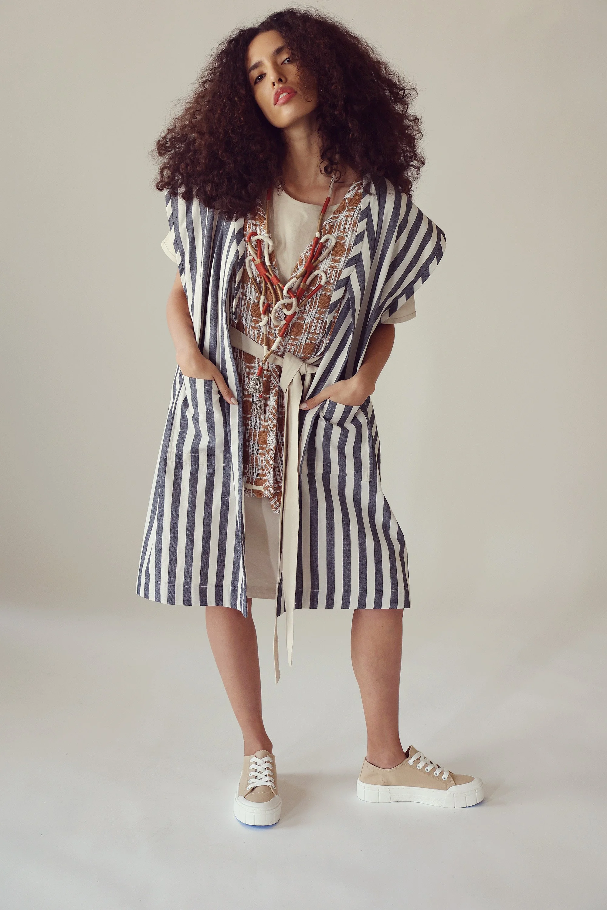 Woman with curly hair wearing a striped sleeveless long coat, layered with a beige shirt and tie-front dress, accessorized with a large beaded necklace, standing against a plain background.