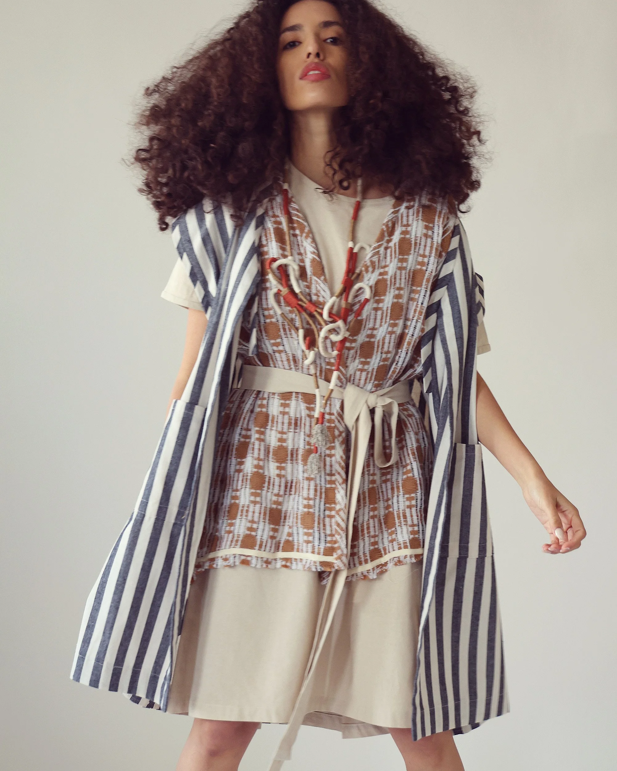 A woman with curly hair wears a layered outfit with a striped coat, patterned vest, and beige skirt, accessorized with a chunky necklace.