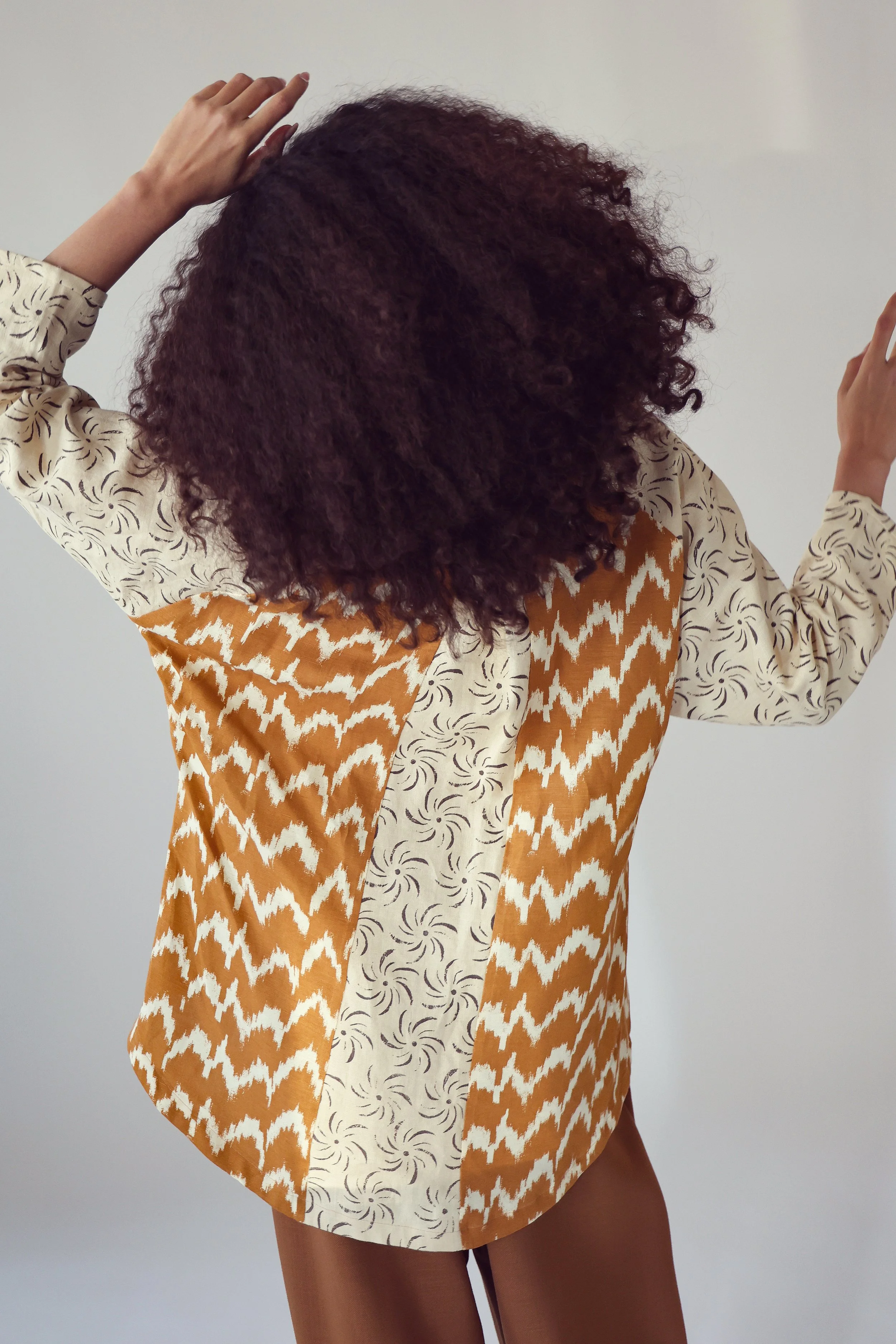 Person with curly dark hair wearing a multicolored patchwork shirt with beige, brown, and cream patterns, standing against a plain white background.