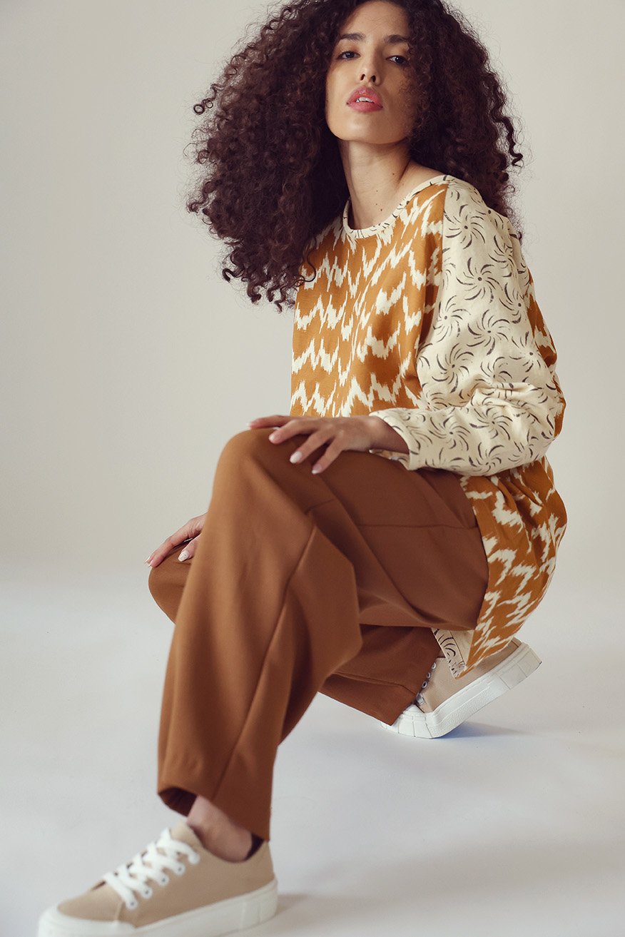 Woman with curly hair wearing a multicolored patterned long-sleeve top, brown wide-leg pants, beige sneakers, sitting on the floor against a plain background.