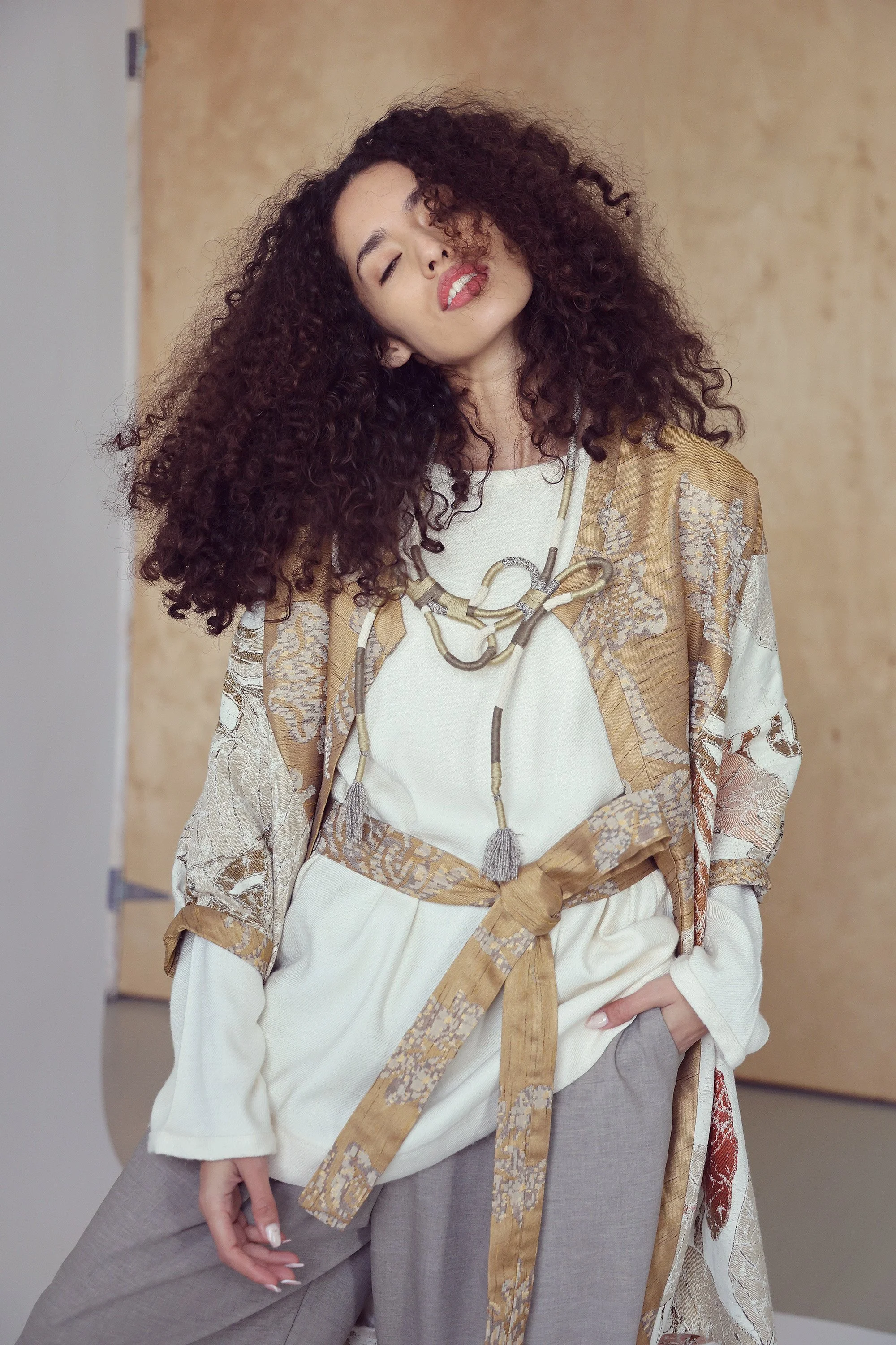 A woman with curly dark hair leaning her head to the side, wearing a patterned kimono-style jacket over a white top, with matching wide-leg pants and a chunky necklace, standing against a beige background.