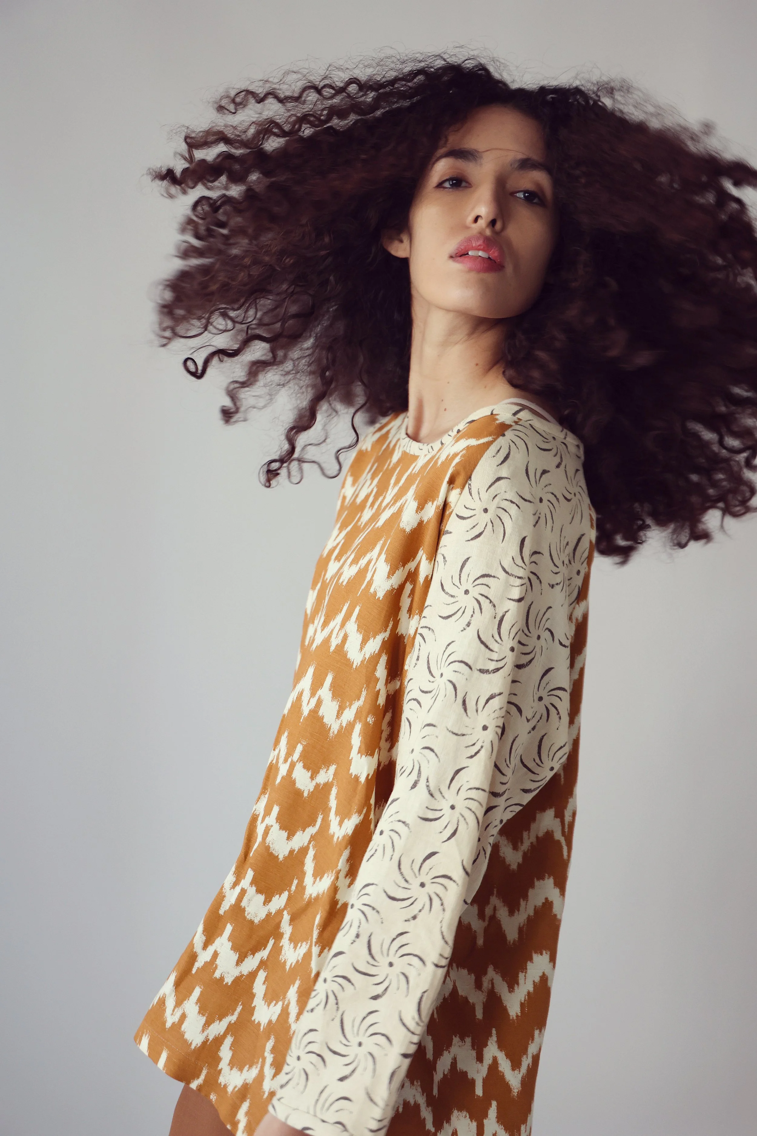 A woman with curly dark hair wears a multicolored patterned dress and stands against a plain background.