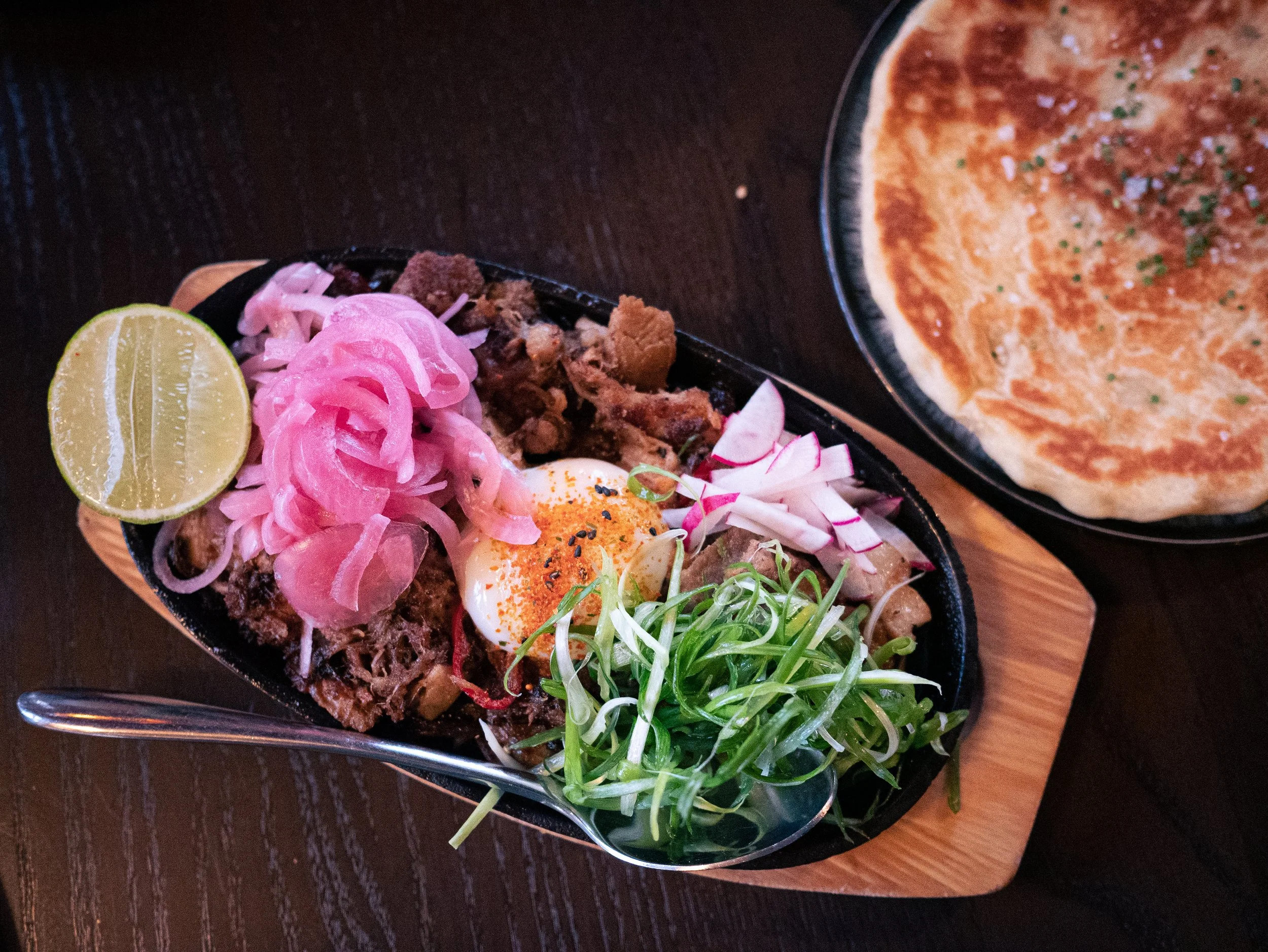 Sising Bing Bread dish from Juju in Durham, NC. Pork buried beneath a soft egg, pickled onions, lime, and other garnishes. Plate of bread to the side.