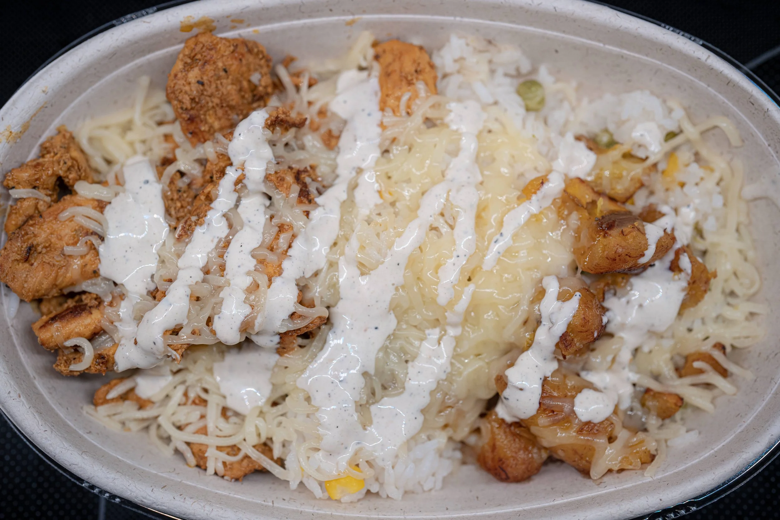 A plate of chicken and rice with cheese, plantains, and garlic sauce. The chicken is cooked in various seasonings. The plantains are baked and sliced, and the garlic sauce is drizzled over the top.