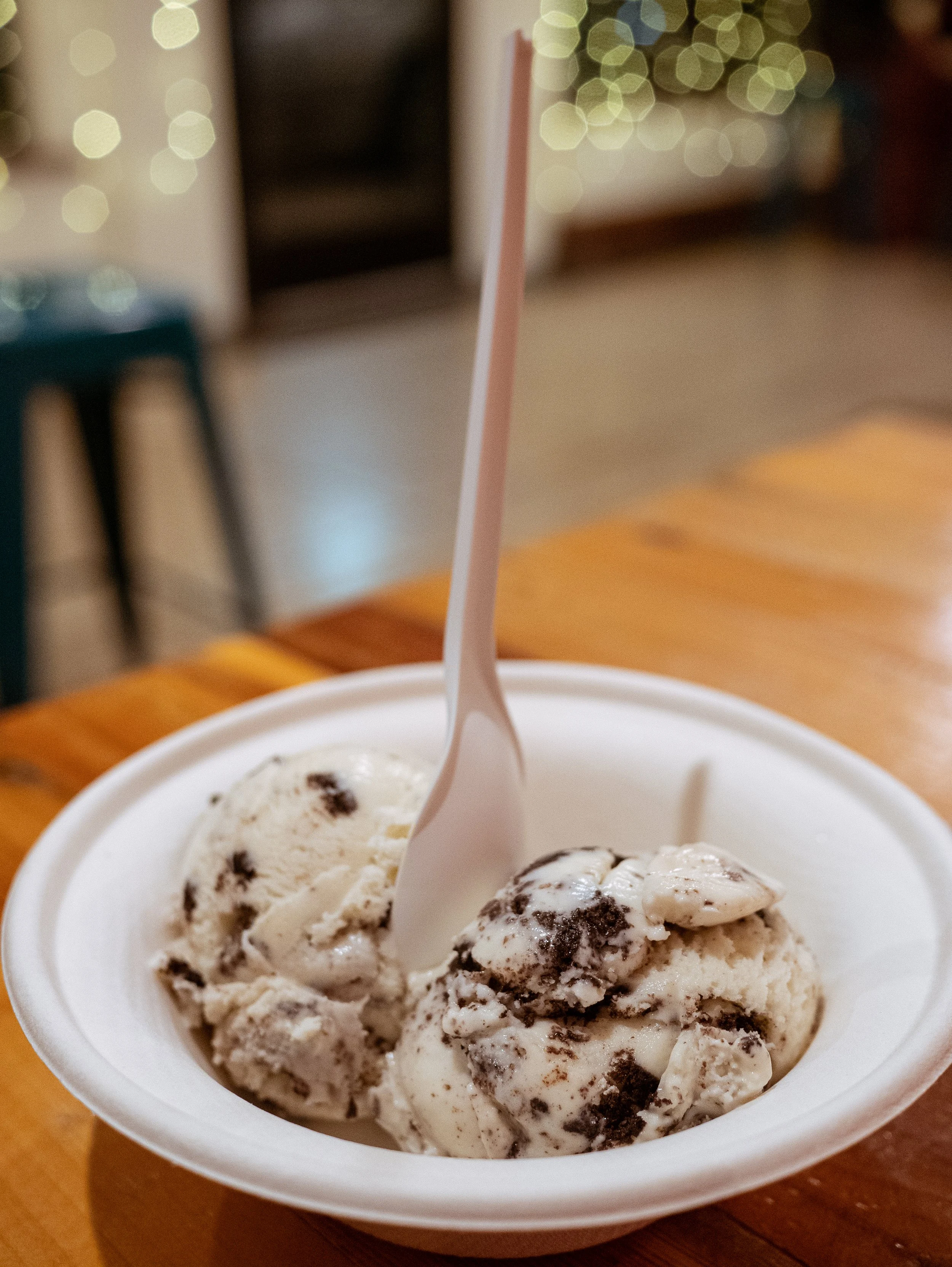 A close-up of 2 scoops of cookies and cream ice cream. The ice cream is creamy and smooth, with chunks of Oreo cookies visible. There are some faint lights in the background.