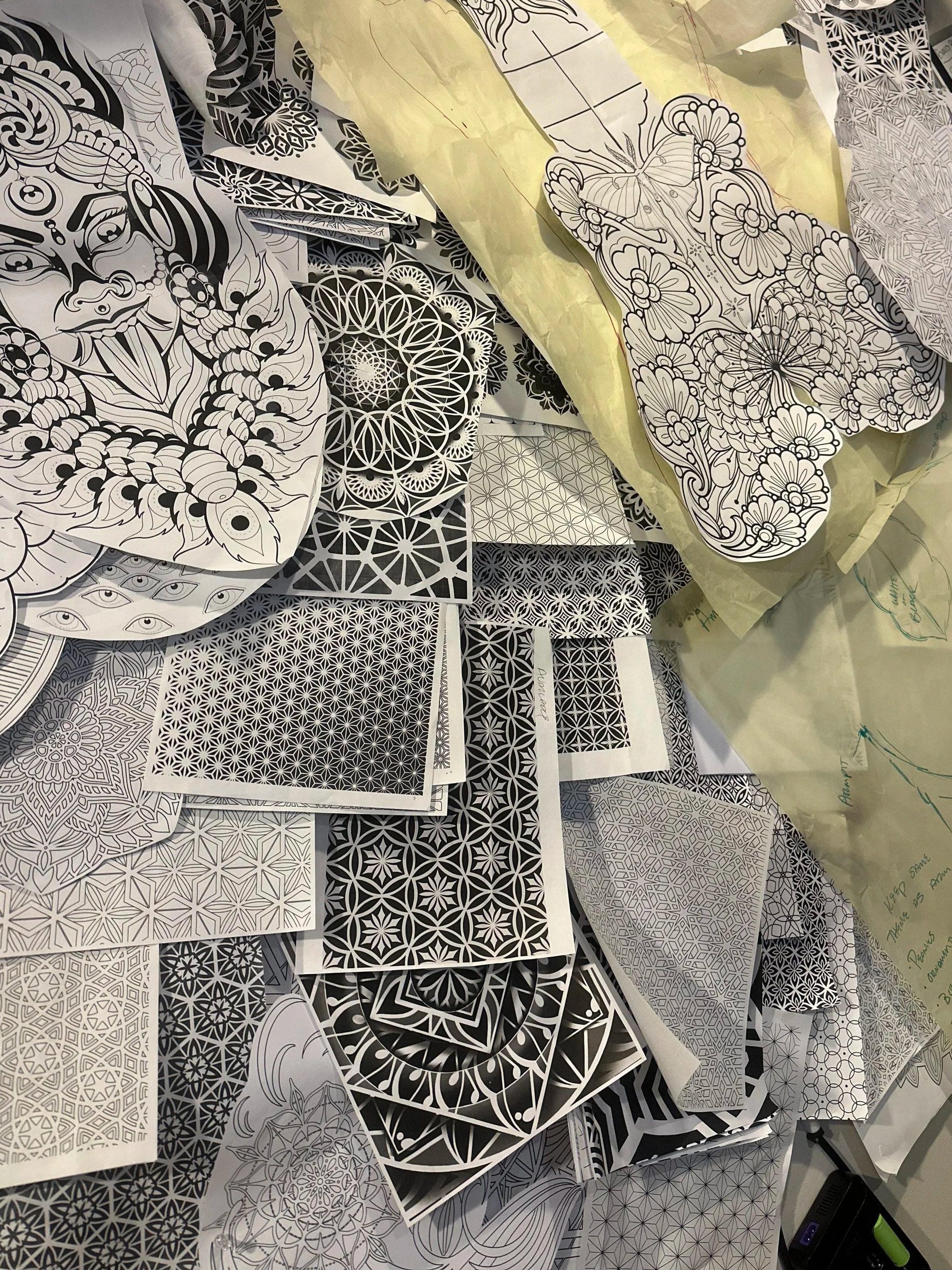 An assortment of numerous black and white intricate mandala and geometric pattern designs, some partially colored, pinned across a cork board.