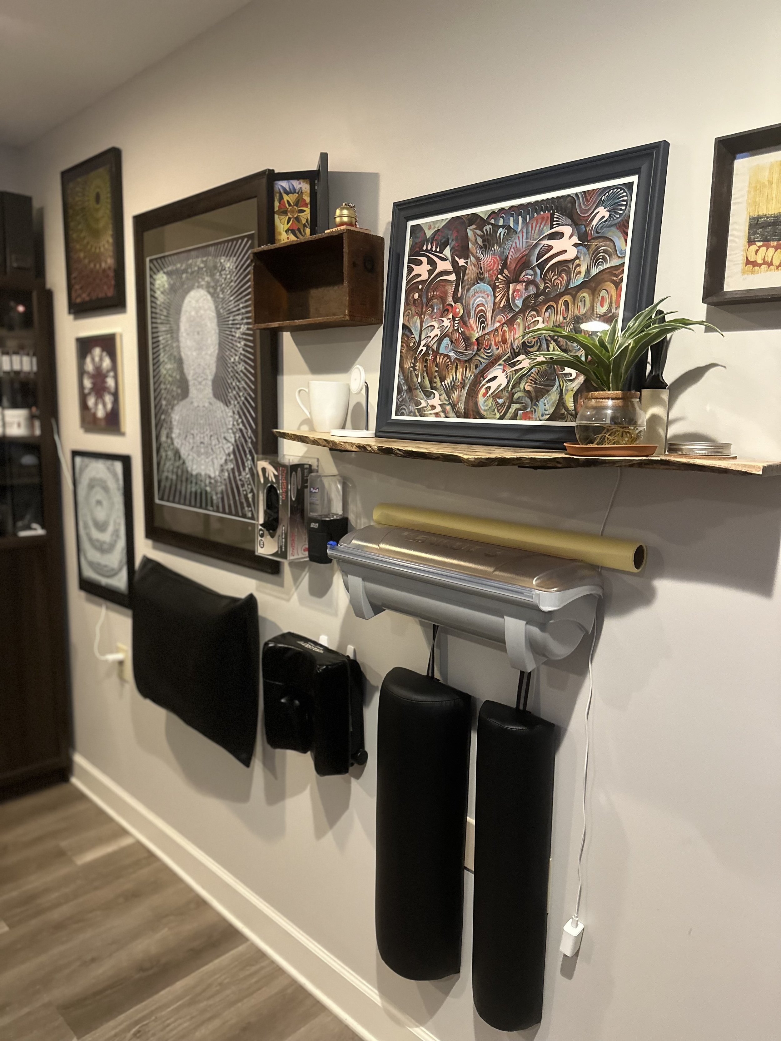 Wall decorated with various framed artwork, a small wooden shelf with decorative items, a potted plant, and black pillows hanging below a wooden shelf.