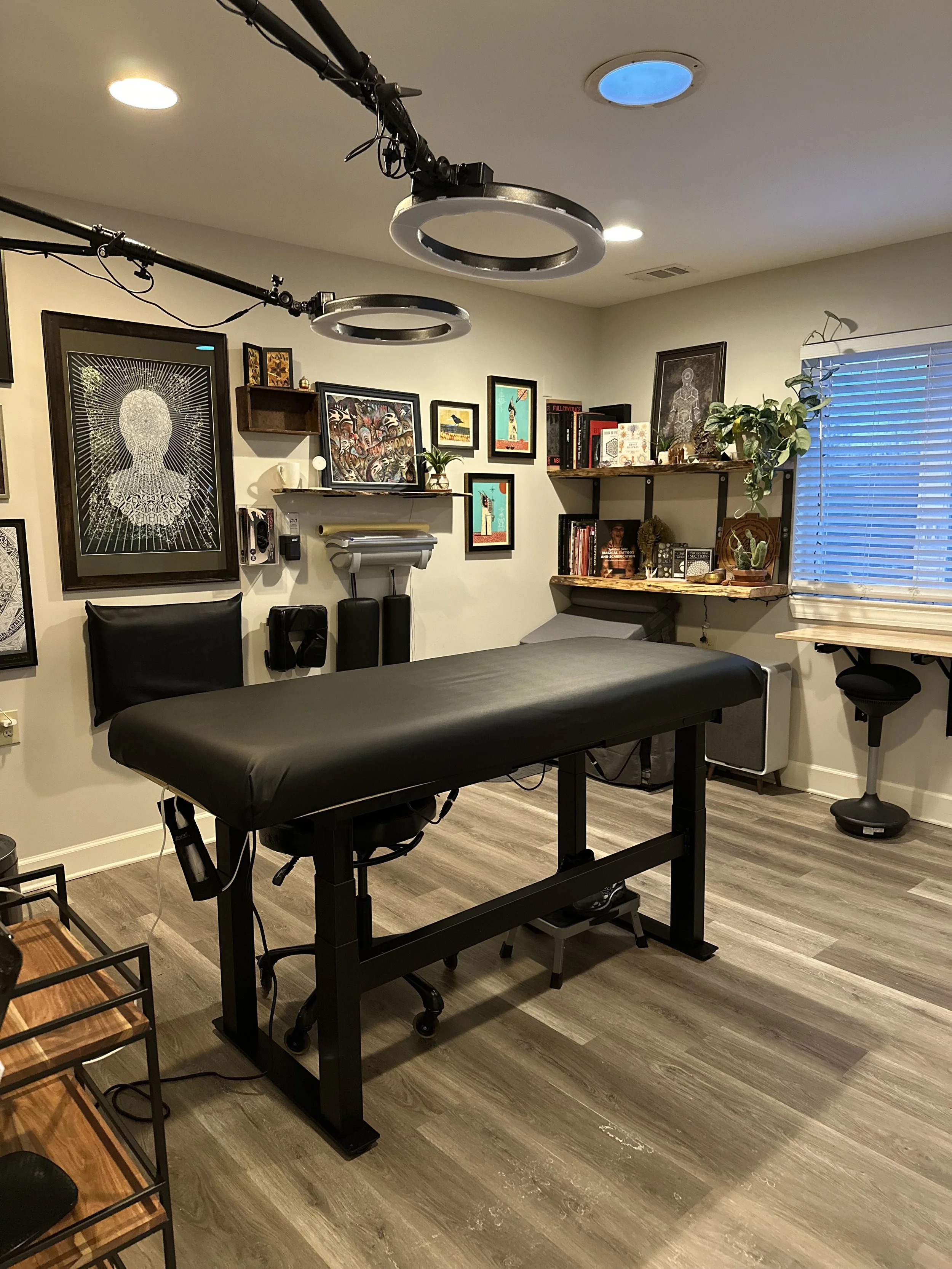 An interior room containing a massage table, office chair, wall art, bookshelves, and chairs with jars, with bright overhead lighting.