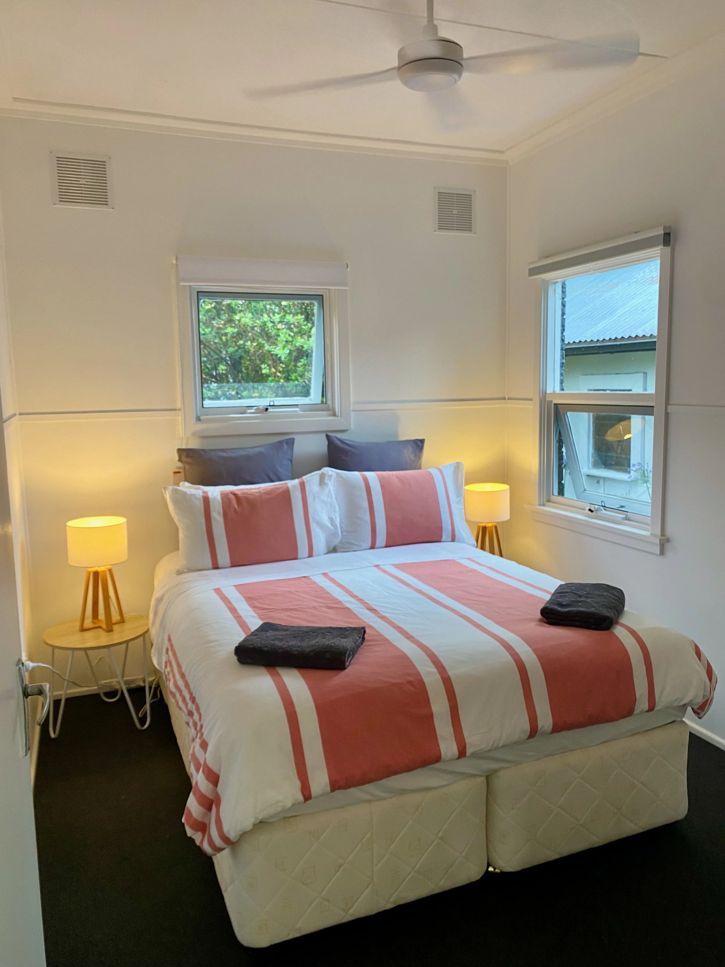 A neatly made bed with pink and white striped bedding and two dark pillows. There are two folded dark towels on the bed. Two small wooden bedside tables with yellow lamps are on either side of the bed. There are two windows, one above the bed and one to the right, letting in natural light. The room has white walls, a ceiling fan, and two ventilation grilles.