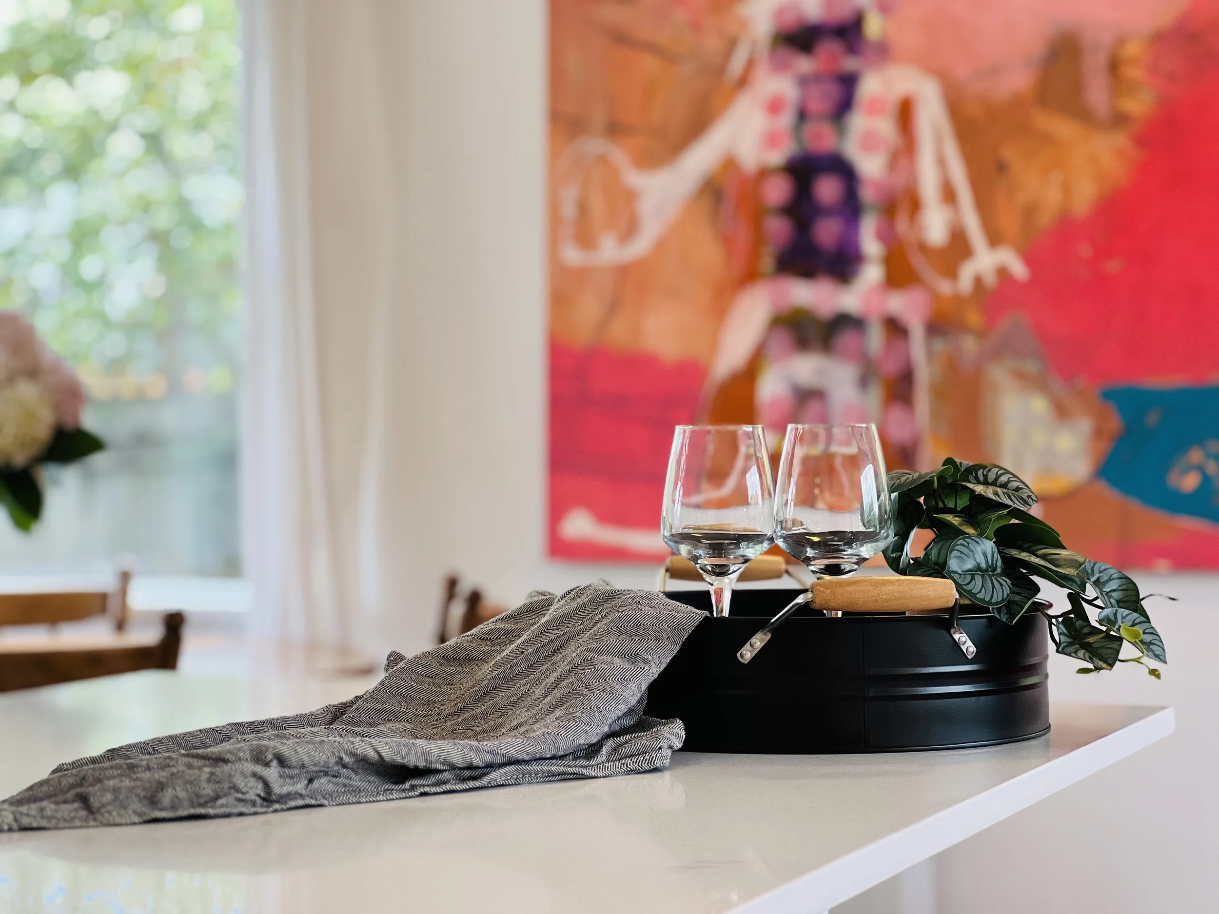 Empty wine glasses on a table with a black planter and green leaves, blurred painting in the background, natural light coming through a window.