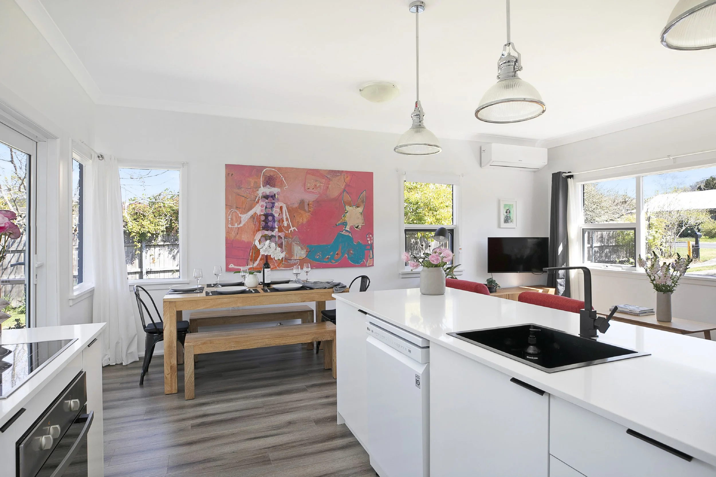 Bright kitchen and dining area with large windows, a table with black chairs, colorful artwork on the wall, and a white island with a black sink.