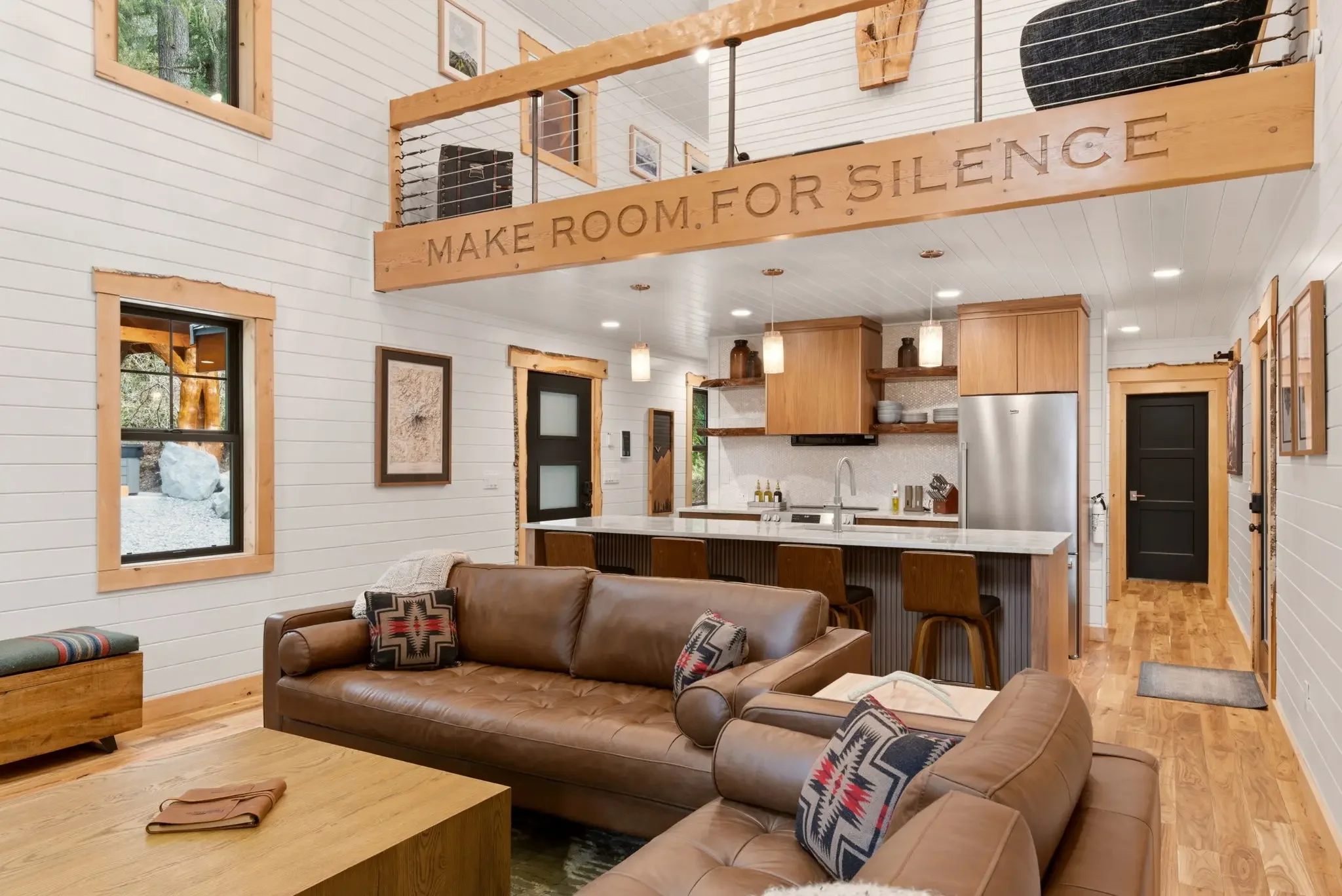 Two-story great room at Fjellsangin cabin near Mount Rainier, with a leather sectional, open kitchen, and a wood loft beam carved with "Make Room. For Silence."