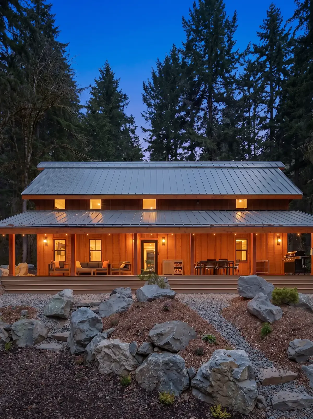 Fjellsangin cabin glowing at dusk with a wraparound porch, metal roof, and string lights, surrounded by evergreens and a stone garden near Mount Rainier.