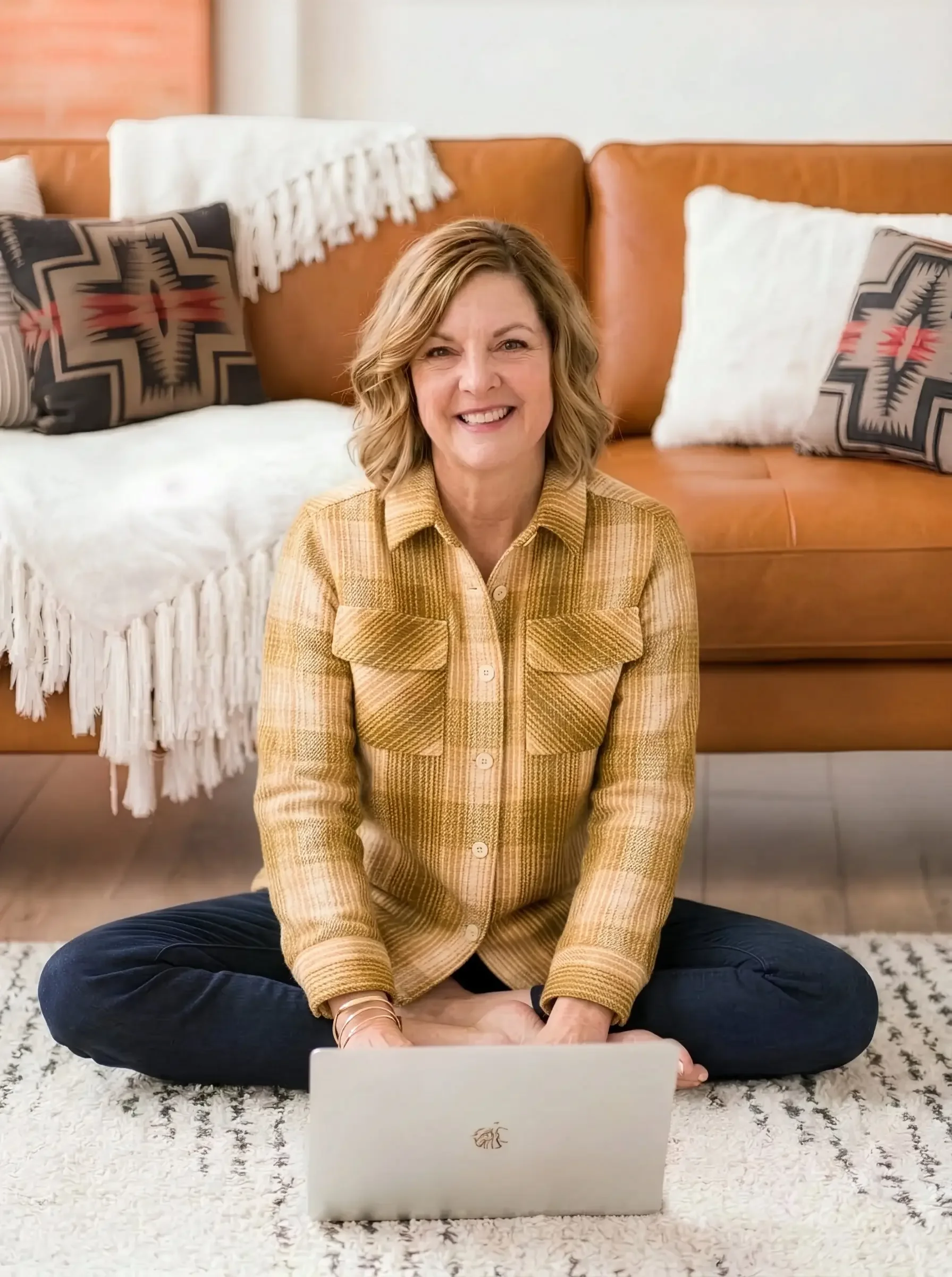 Jennifer Mager, founder of Nest & Nook Creative, sitting cross-legged on a rug with a laptop, smiling in front of a leather sofa with patterned pillows.