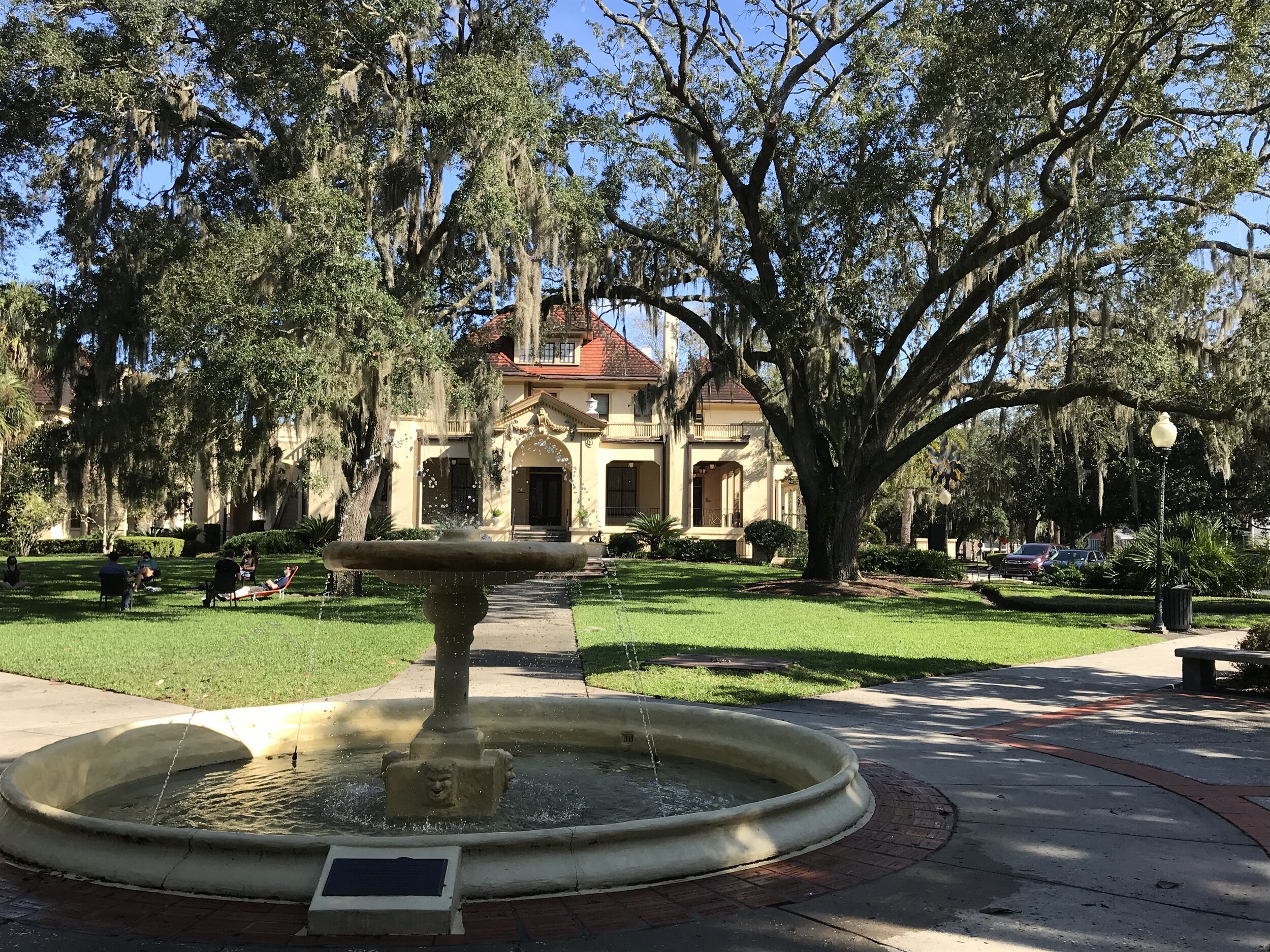 A serene park scene featuring a round stone fountain with water spouts in the foreground, large oak trees draped with Spanish moss, and a historic building in the background. People are relaxing on the green grass, enjoying the sunny day.