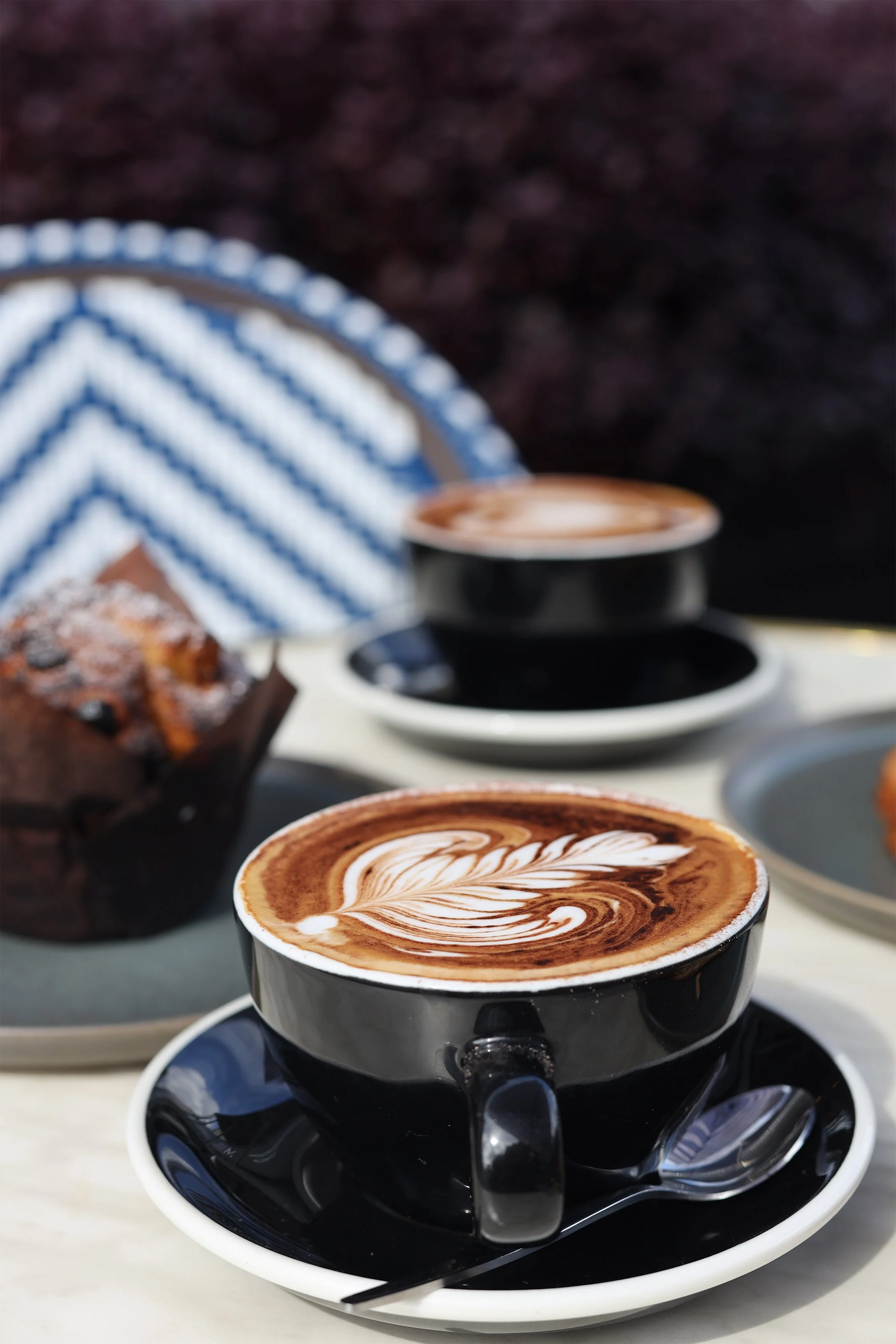 Two cups of cappuccino with latte art, a chocolate muffin, and a spoon on a table outdoors.