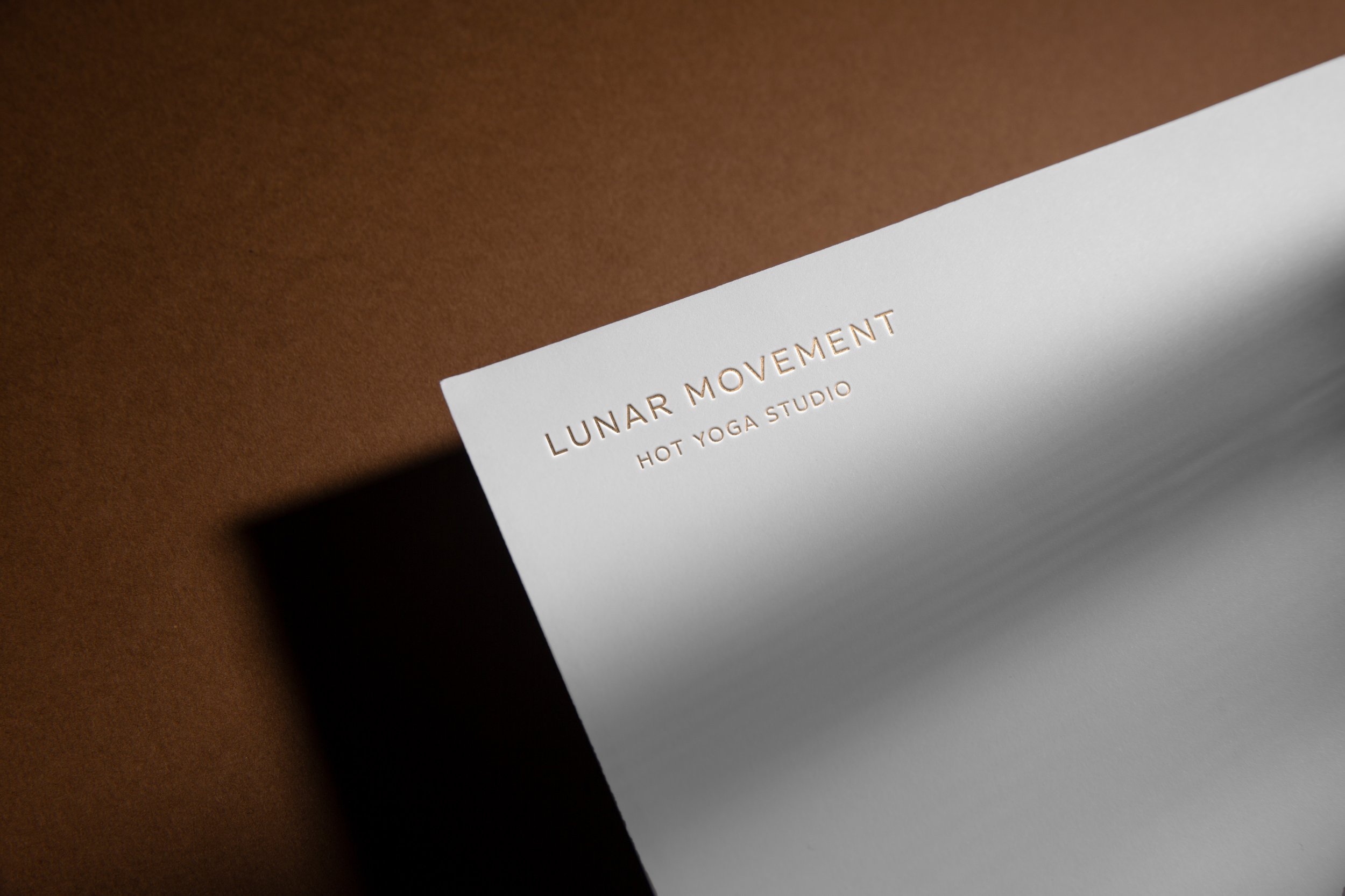 Close-up of a white paper with gold embossed text reading 'LUNAR MOVEMENT HOT YOGA STUDIO' on brown surface.