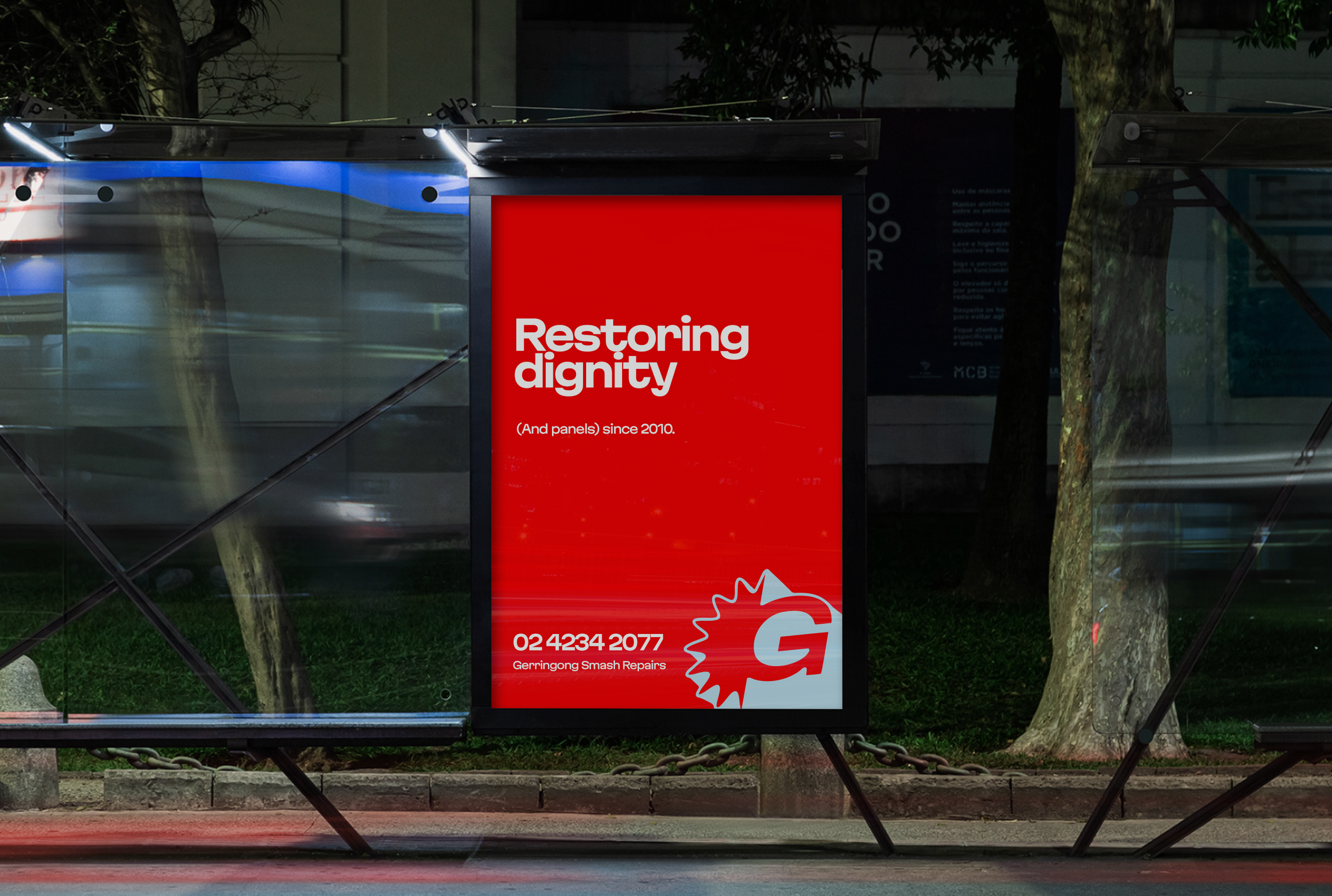 Red advertising poster on a bus stop at night, with text about restoring dignity and a phone number, featuring a Gerringong Smash Repairs logo, surrounded by trees and reflections of passing vehicles.