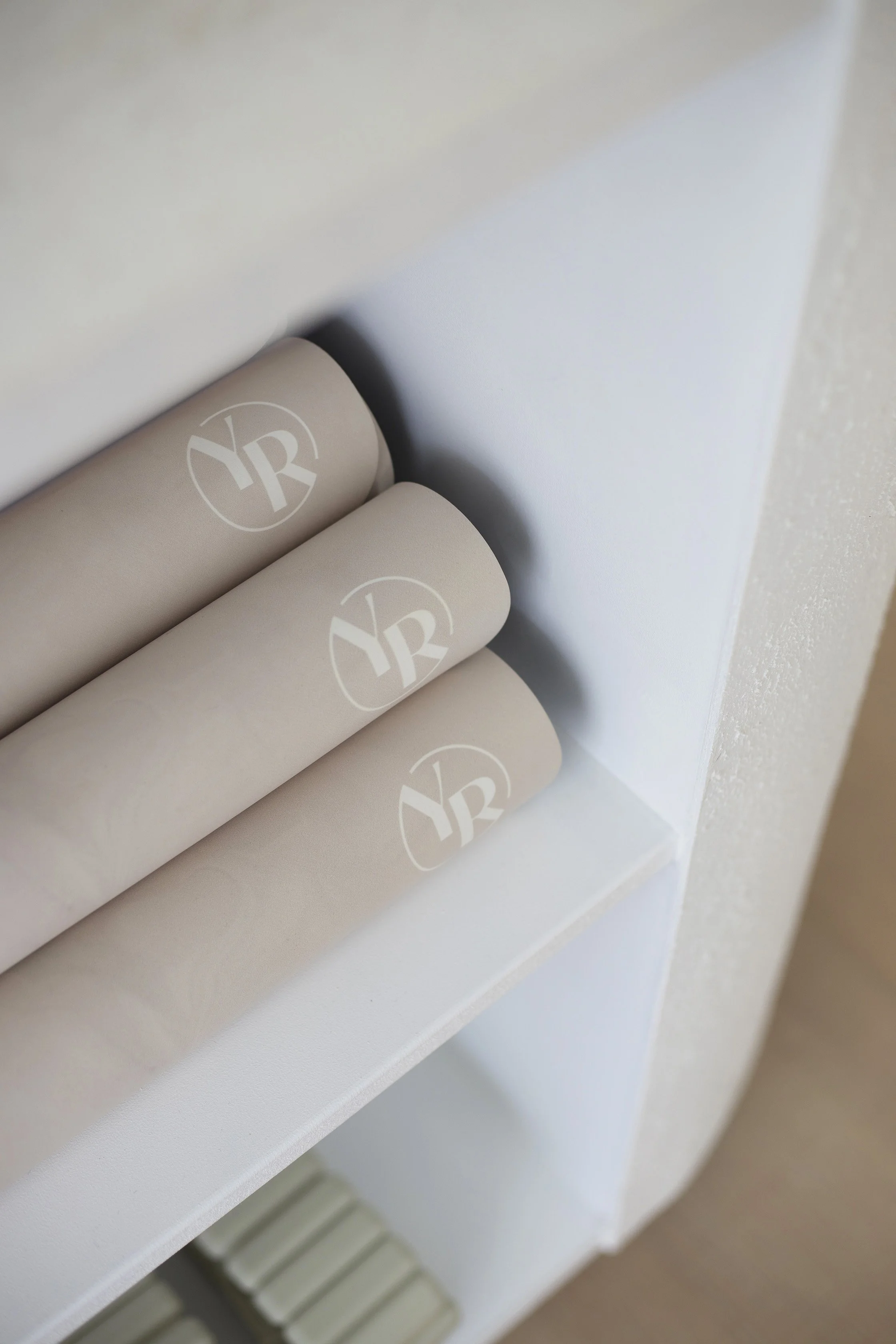 Three beige cylindrical candles with white logos on a white shelf, behind a white wall.