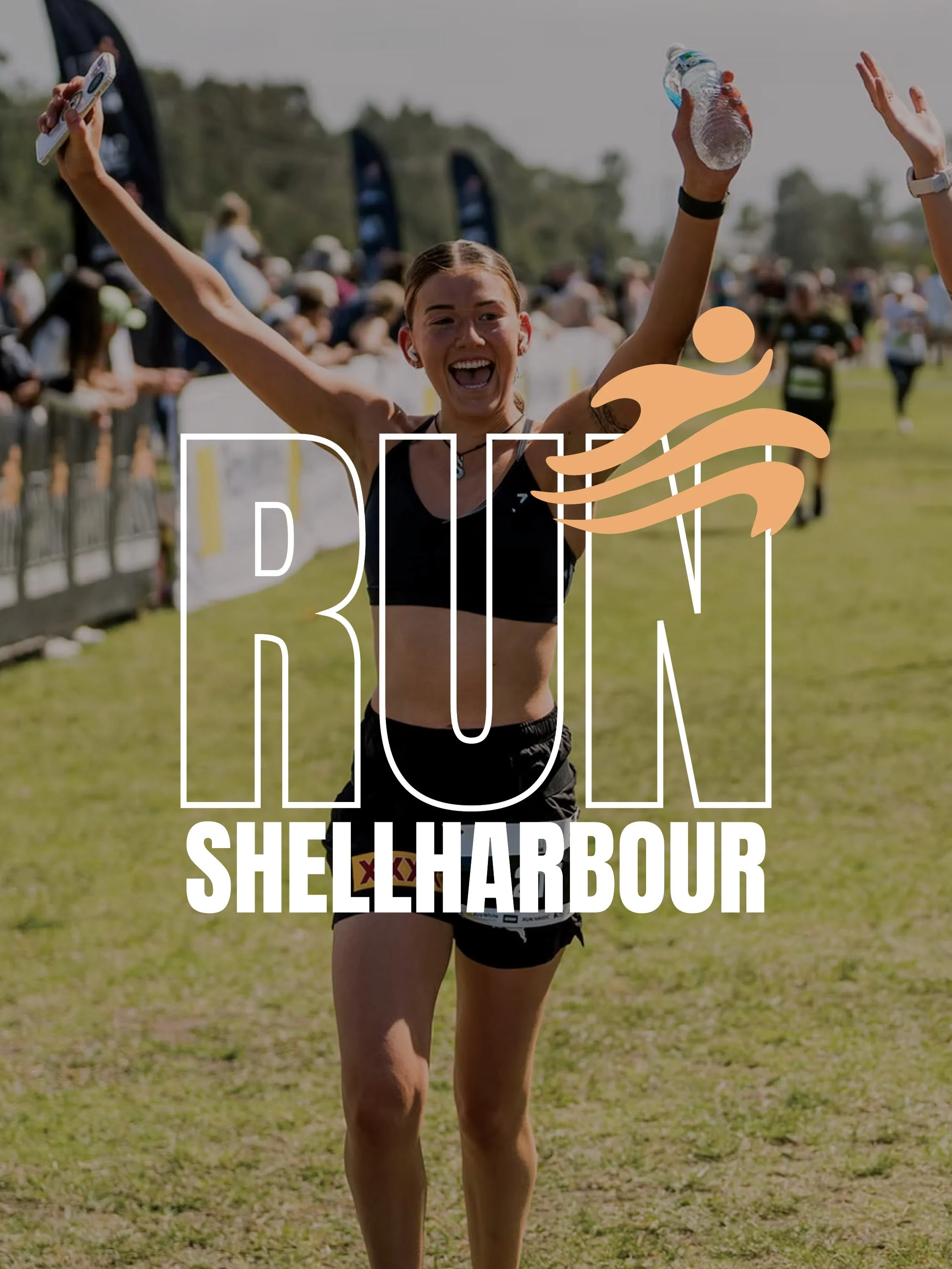 A female runner celebrating crossing the finish line at Shellharbour event, with arms raised, holding a water bottle and a medal, crowd cheering in the background.