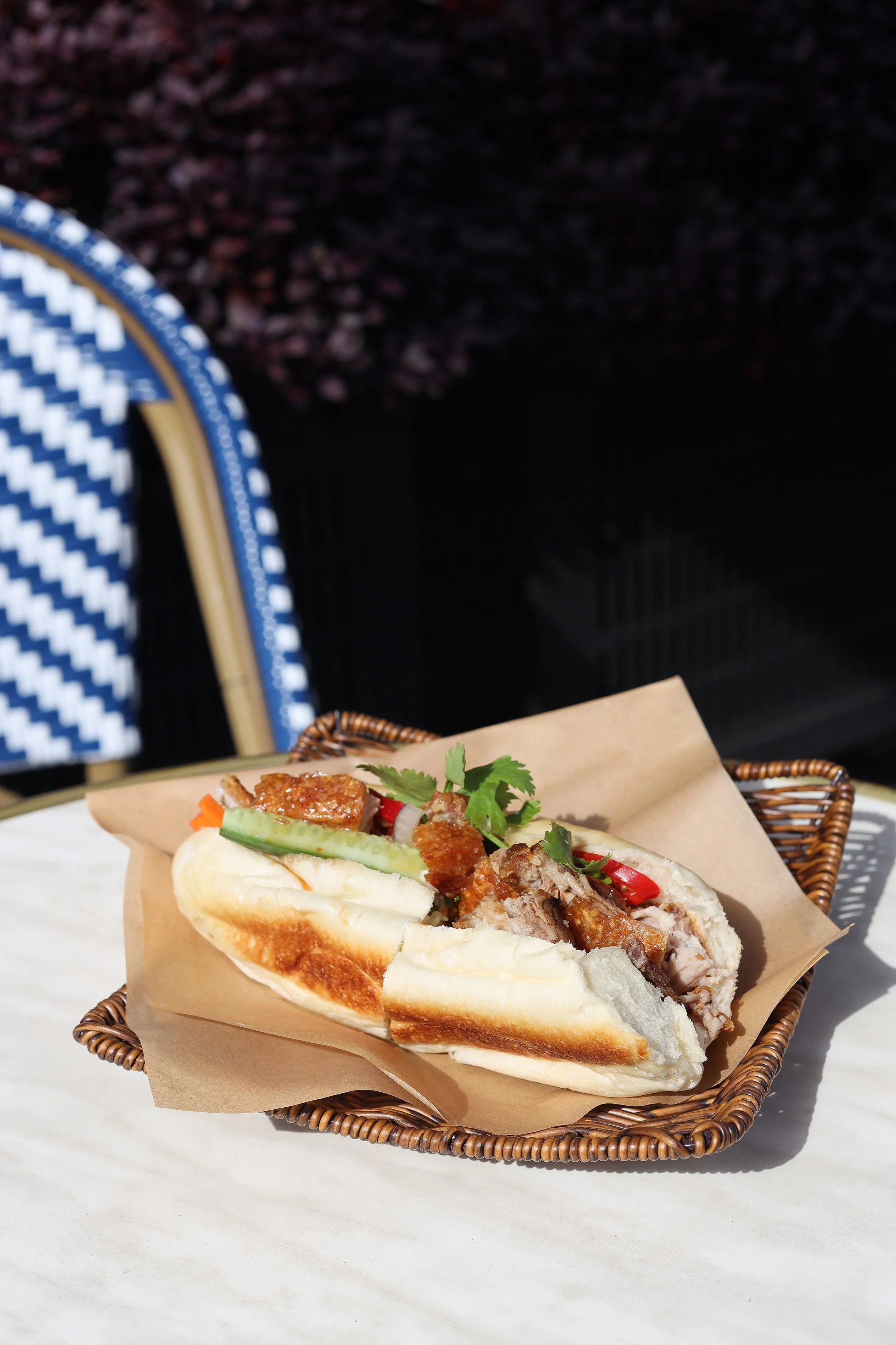 Vietnamese banh mi sandwich in a small basket with pickled vegetables, cilantro, cucumber, and grilled meat on a white table outside.