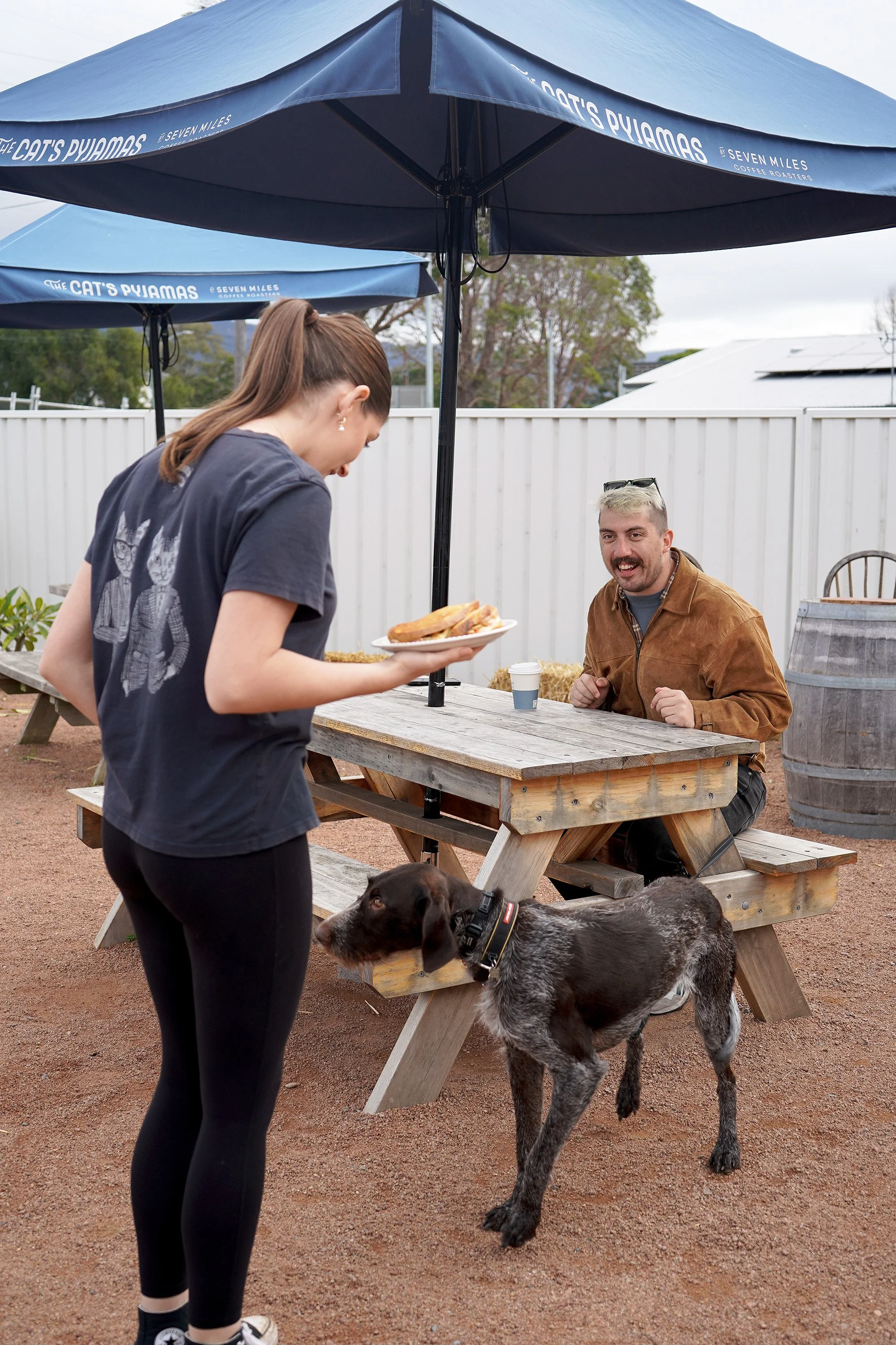 A young woman holding a plate of food and a dog looking at her, while an older man sits at a picnic table under a large blue umbrella, smiling.