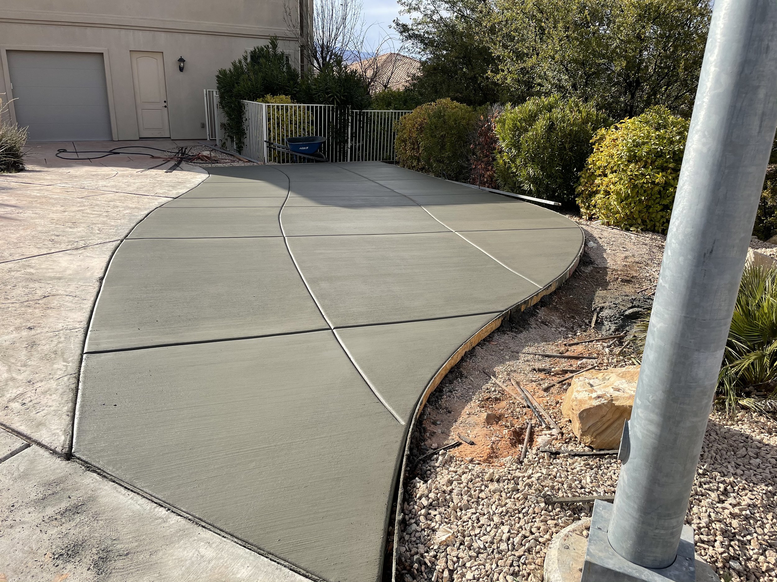 Photo of a new concrete sidewalk with curved edges next to a landscaped area with bushes and rocks, a white metal pole, and a white building with a garage door and a door with a light fixture.