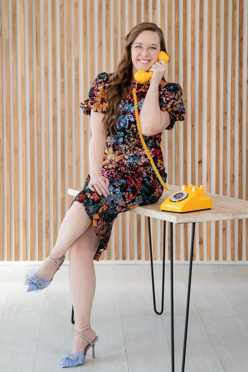 Madigan, founder of CommitmentISM, smiles while holding a yellow vintage phone, conveying warmth, approachability, and an invitation to connect.