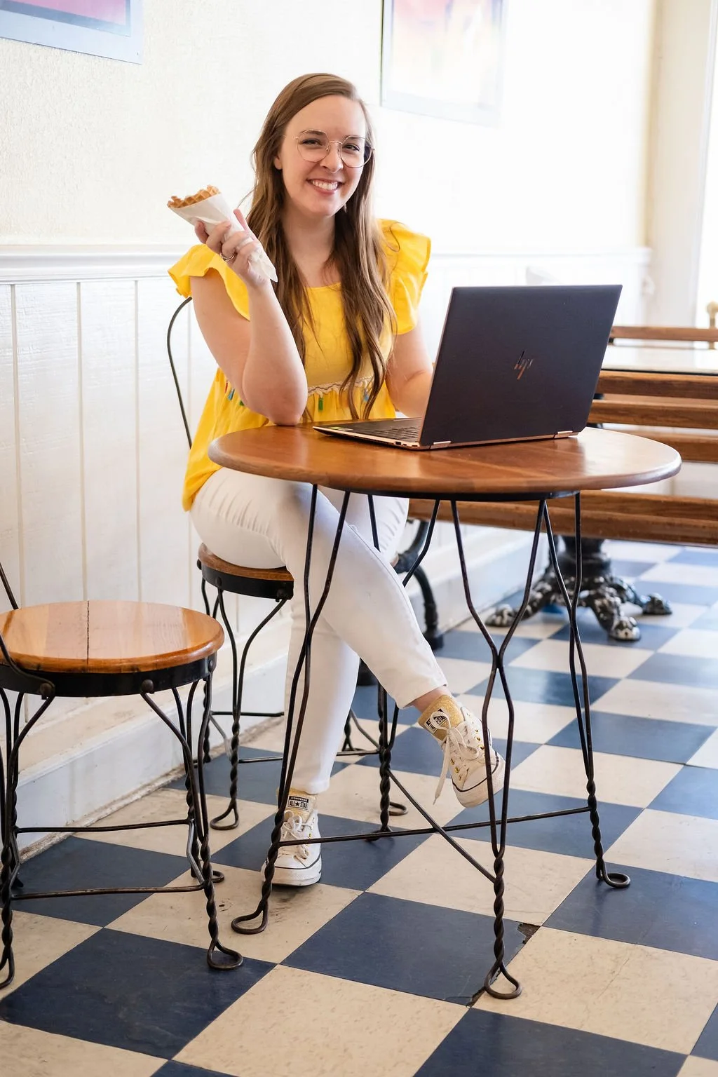 Madigan, founder of CommitmentISM, smiles while seated at a café table with a laptop and pastry, conveying warmth, approachability, and everyday relatability