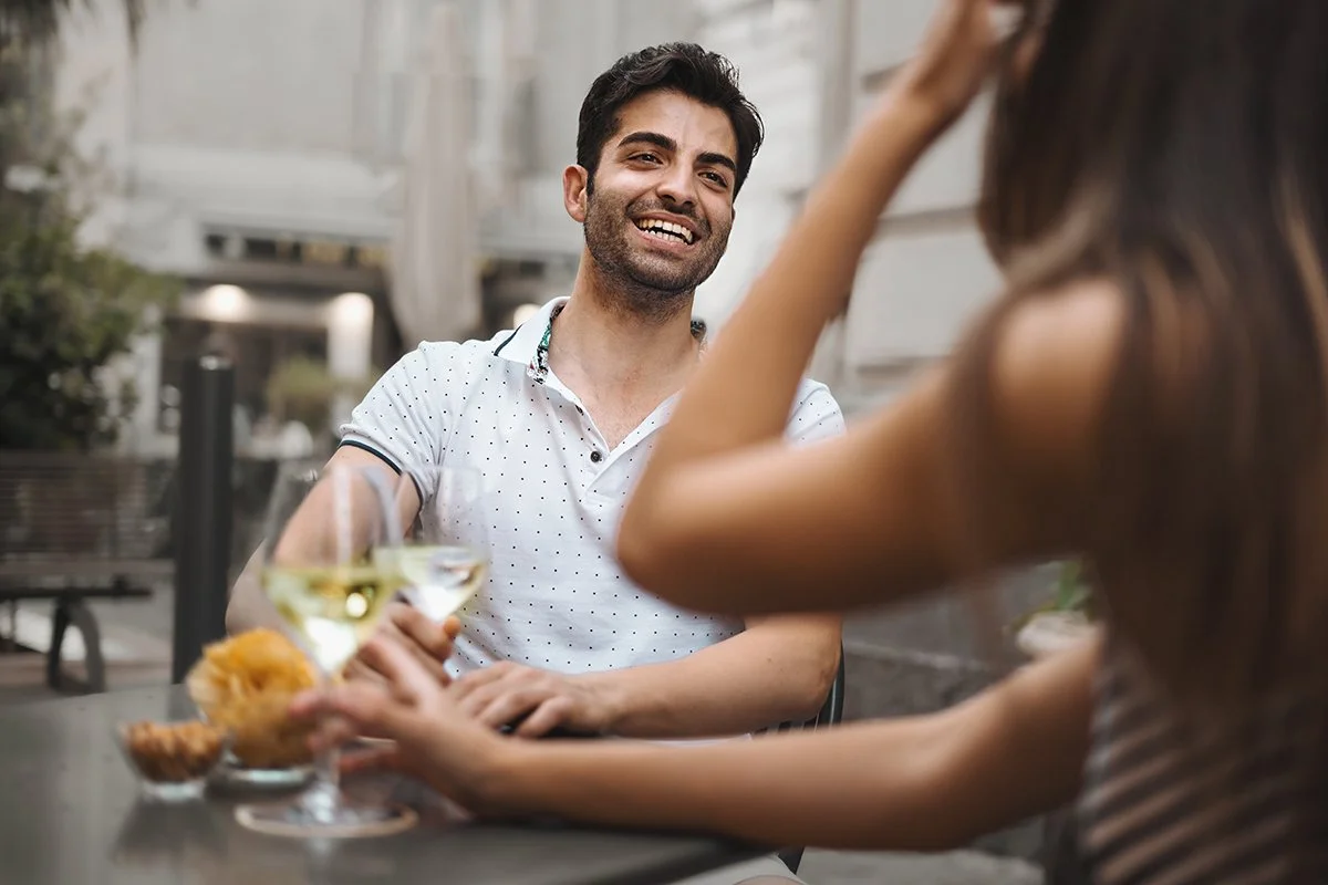 A man smiles while engaged in relaxed conversation over drinks, reflecting ease, interest, and genuine connection.