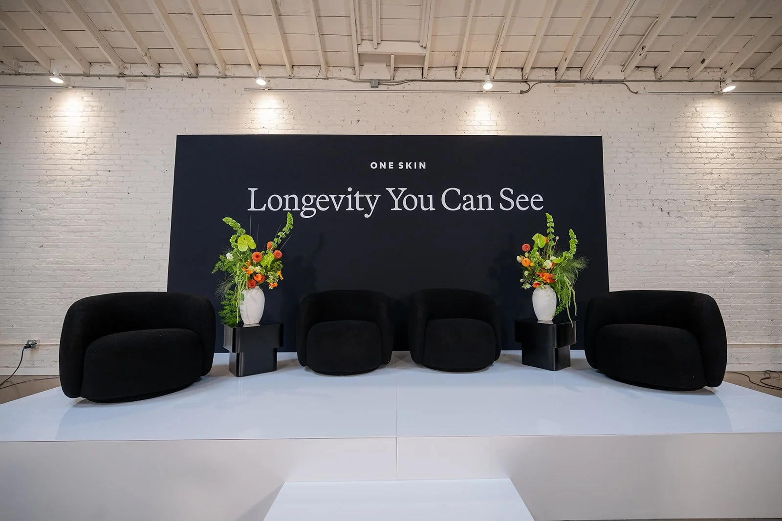Minimalist panel stage with black lounge chairs and “Longevity You Can See” branding at OneSkin event