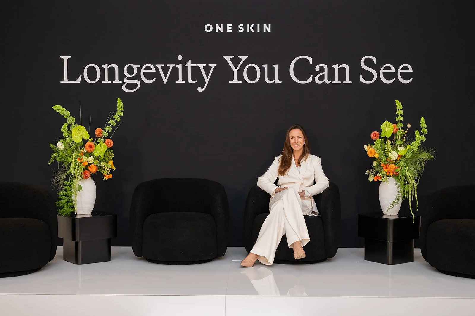 Minimalist panel stage with black lounge chairs and “Longevity You Can See” branding at OneSkin event