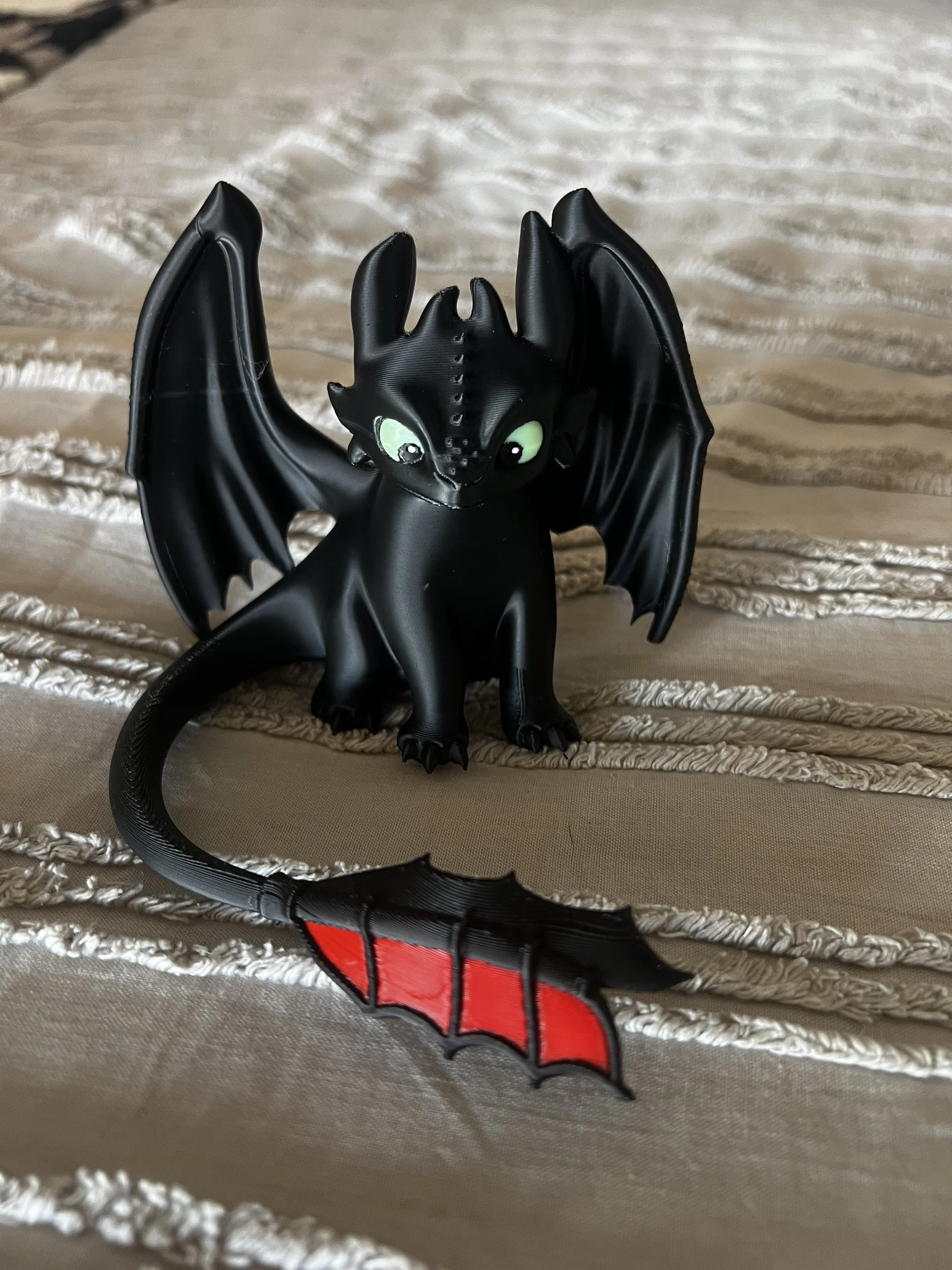 A black figurine of Toothless, the dragon from How to Train Your Dragon, with green eyes and wings spread out, standing on a textured surface. In the foreground, part of a red and black figure resembling Spider-Man's mask is visible.