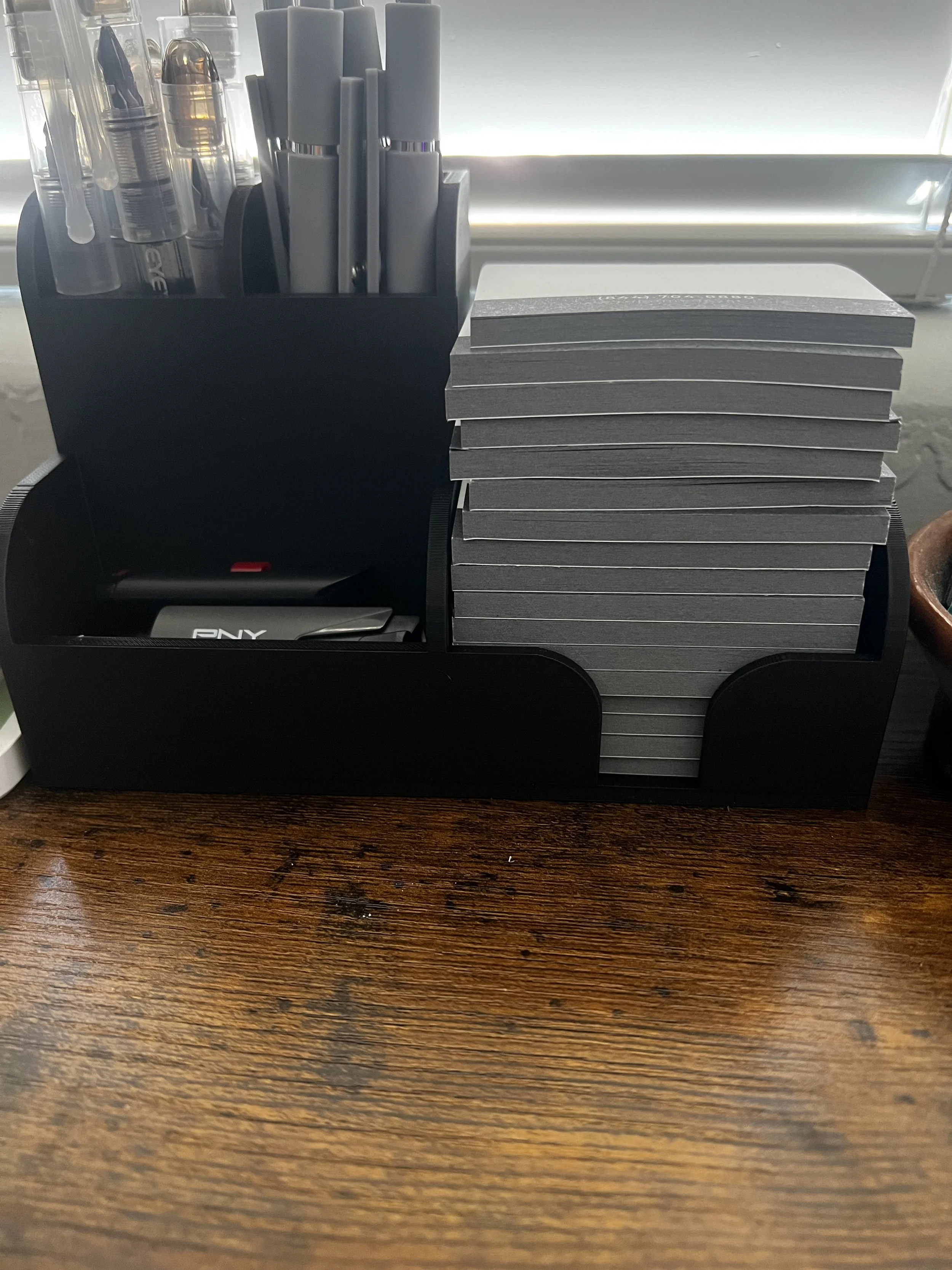 A black desktop organizer holding pens, highlighters, and a pack of Post-it notes on a wooden table.