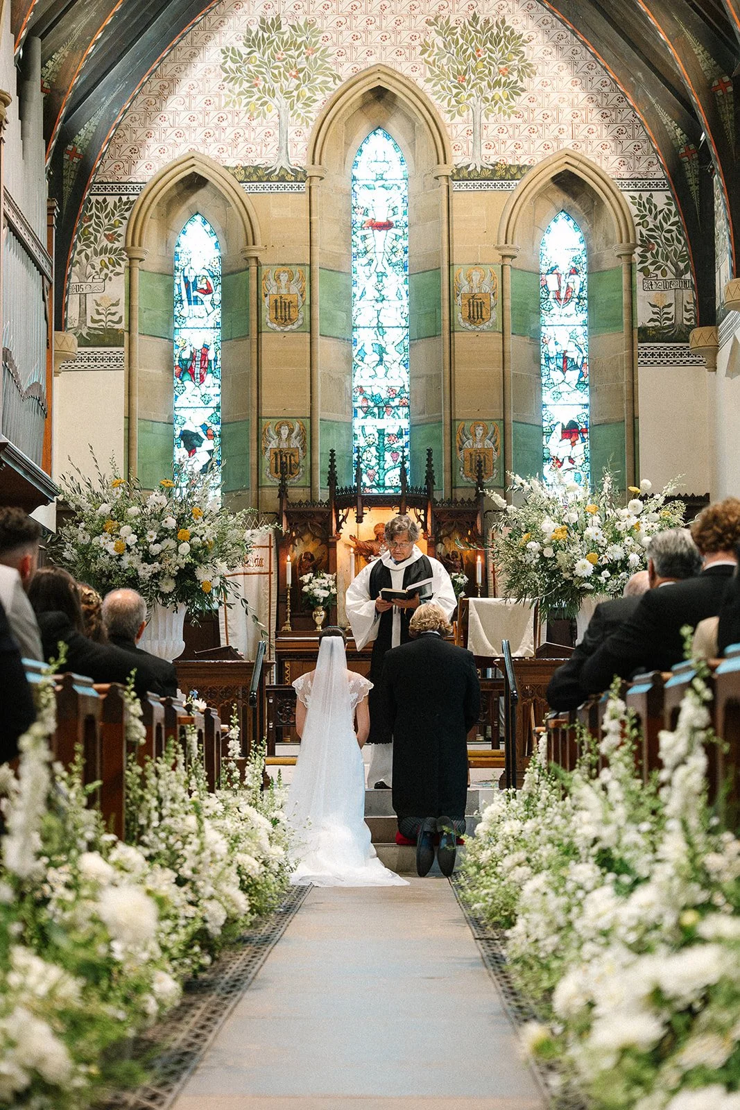 Bride and groom kneel at the church altar framed by two abundant floral urn arrangements, the aisle is lined with flowers