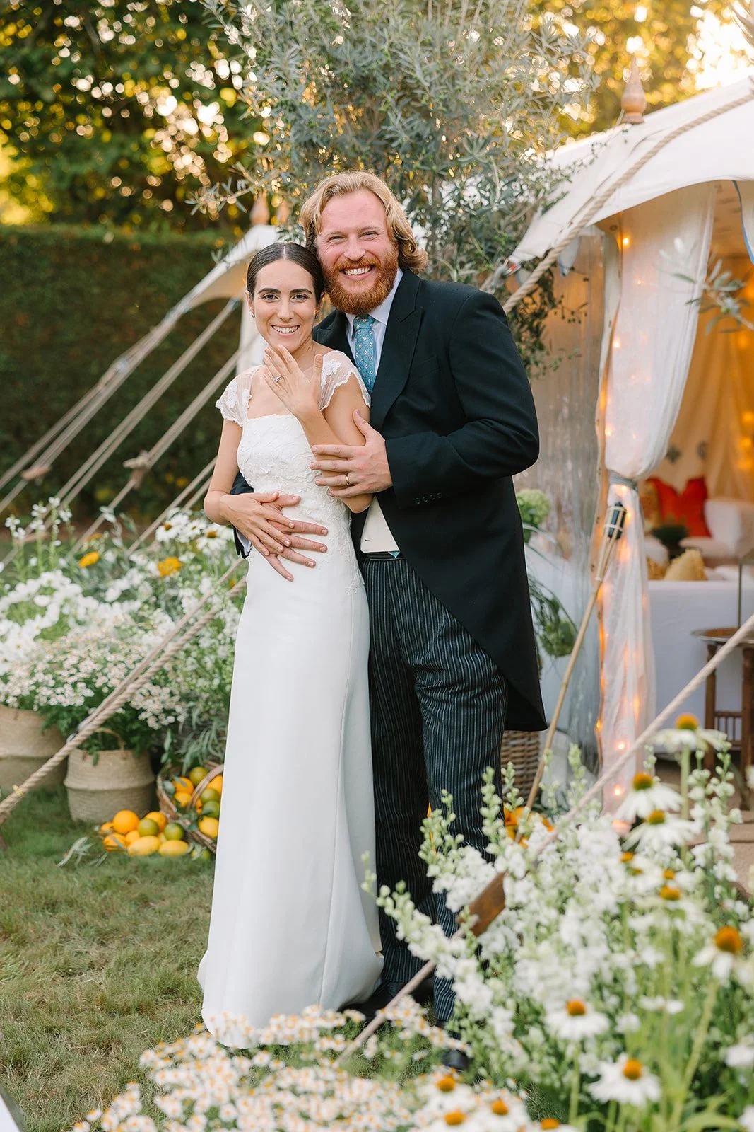 Bride and groom at the entrance to a traditional sail cloth marquee, the bride wears a traditional lace wedding dress and teh groom weras morning dress. The netrance to the marquee is decorated with wicker baskets of flowers and olive trees