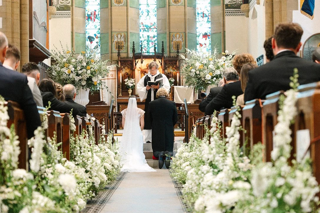 Bride and groom kneel at the altar to take their wedding vows. The aisle is lined with flowers and two large floral urn arrangements frame the couple