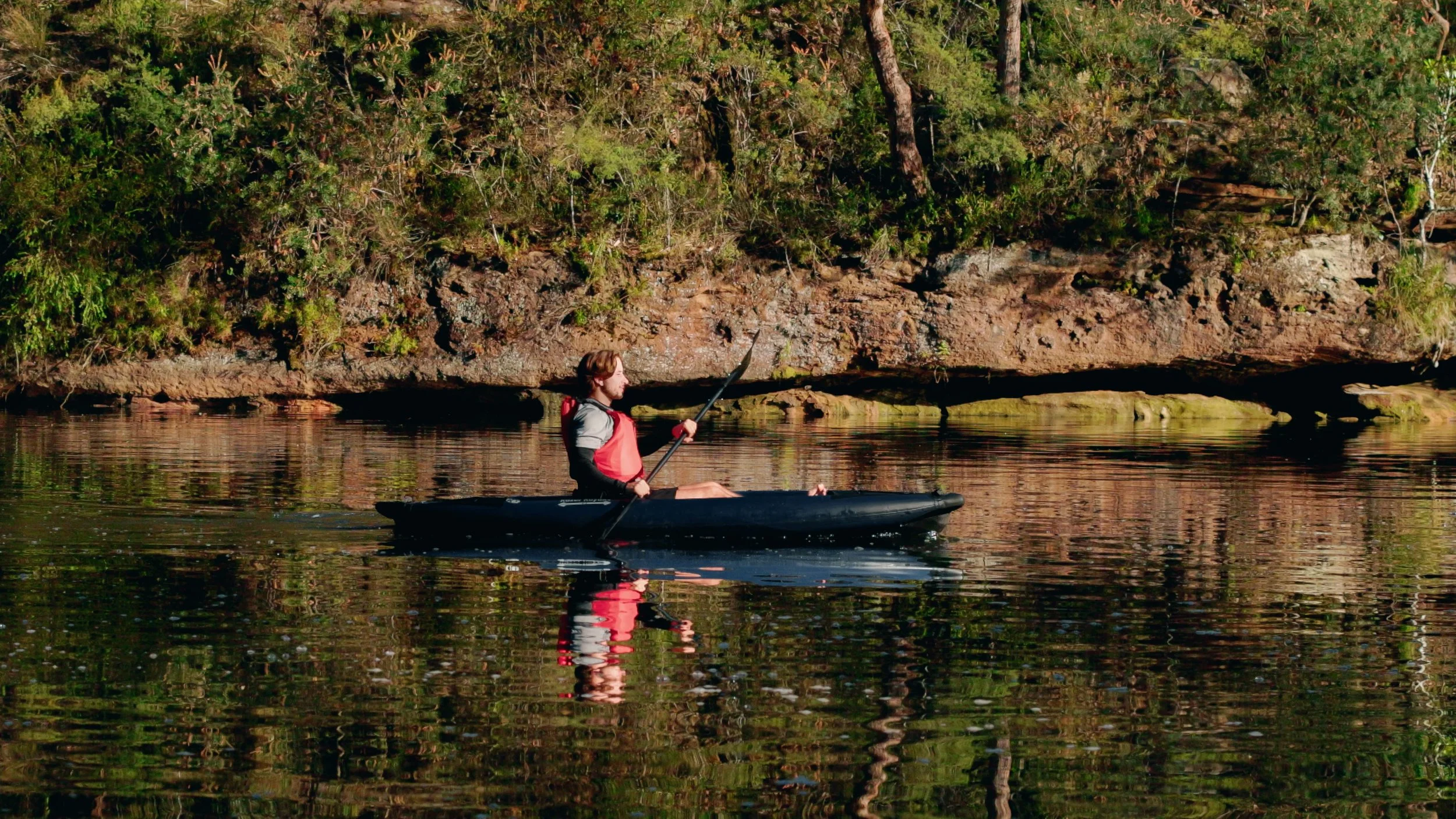 Pagayeur dans un kayak gonflable Razor Kayaks R1 Ultra sur une eau calme près d’une rive boisée.
