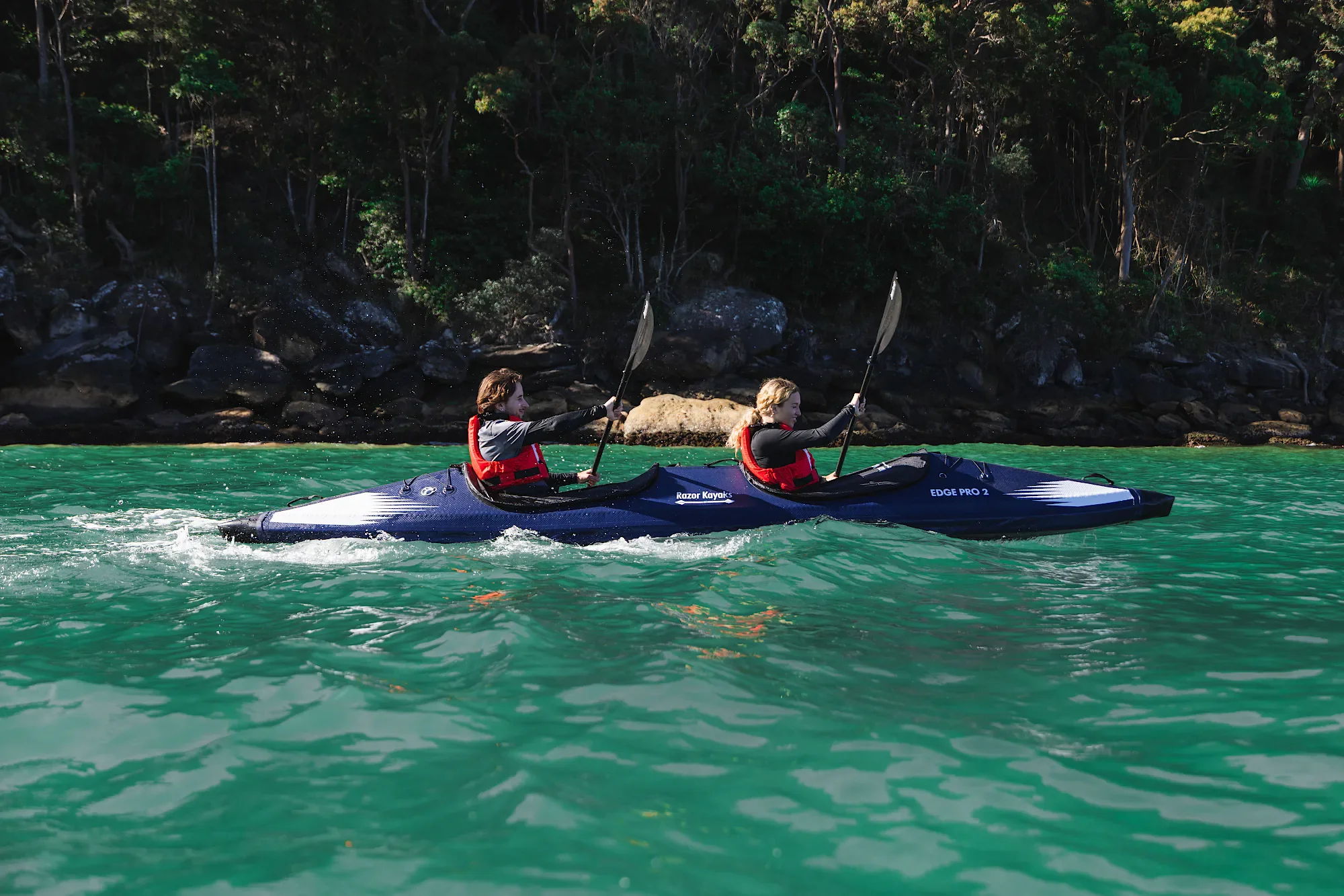 Deux pagayeurs dans un kayak gonflable Razor Kayaks Edge Pro 2 près d’une rive rocheuse et boisée.