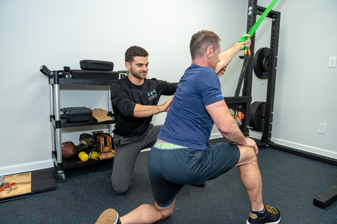 A physical therapist assists a man doing a stretching exercise with resistance bands in a gym.