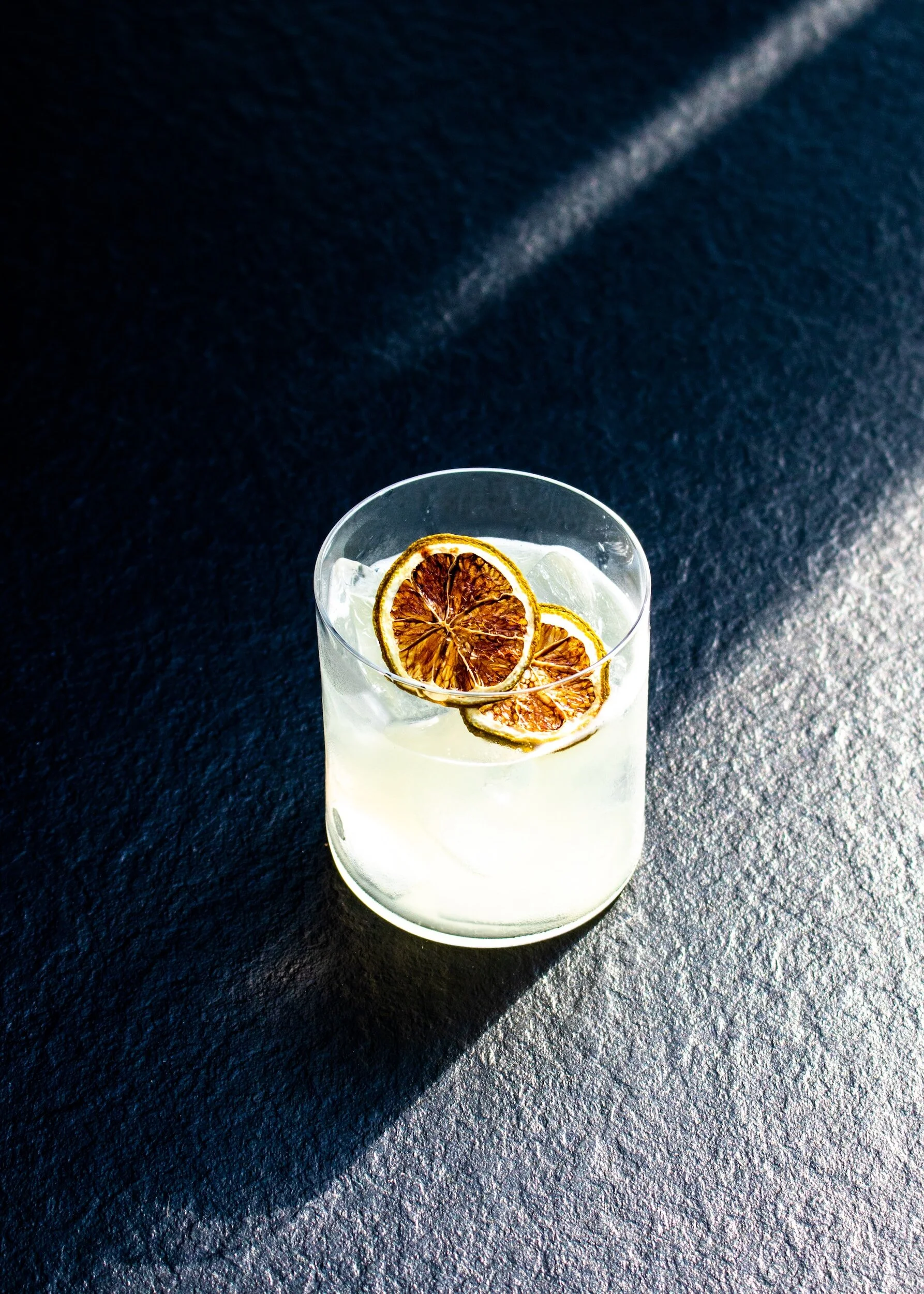 A clear glass of lemonade with ice cubes and slices of dried lemon on top, placed on a dark textured surface with a shadow cast to the left.