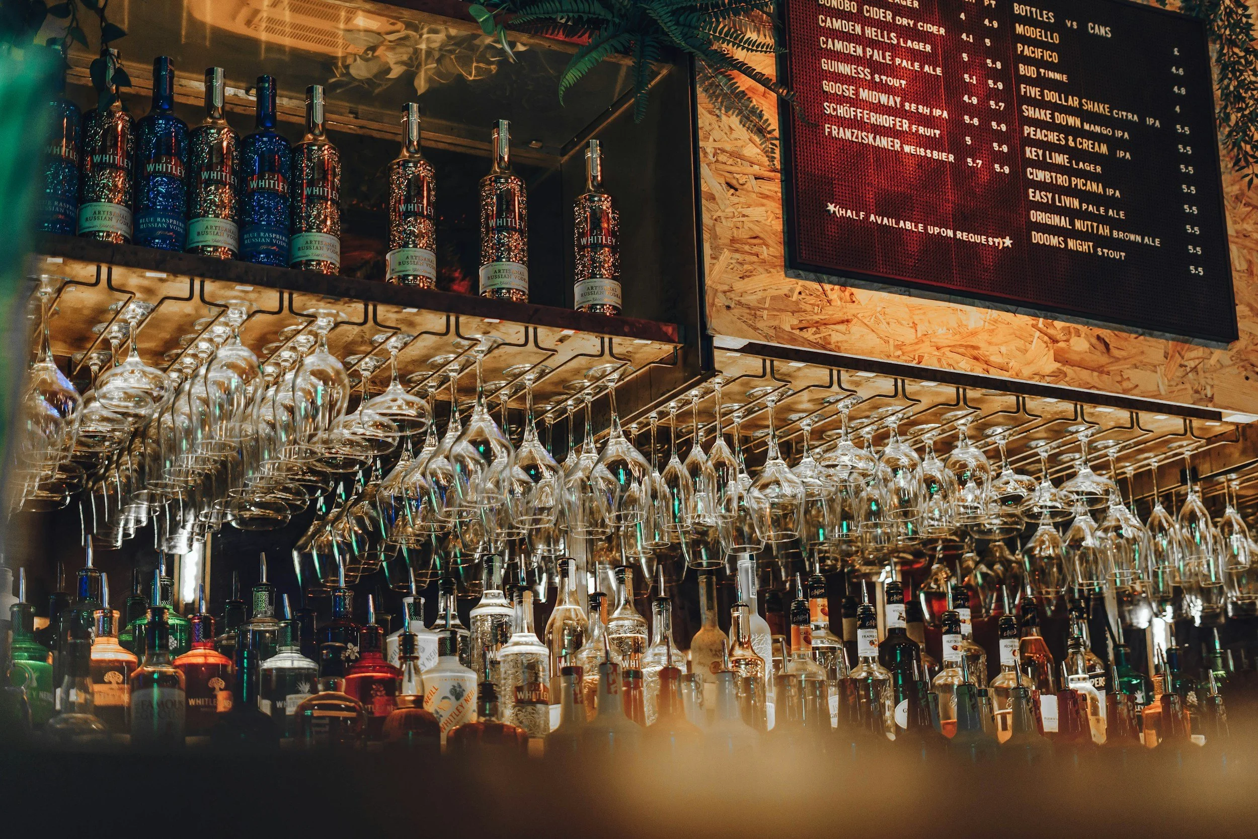 Bar shelf displaying wine and liquor bottles with hanging wine glasses underneath, and a red digital menu board with drink names and prices on a wooden wall.