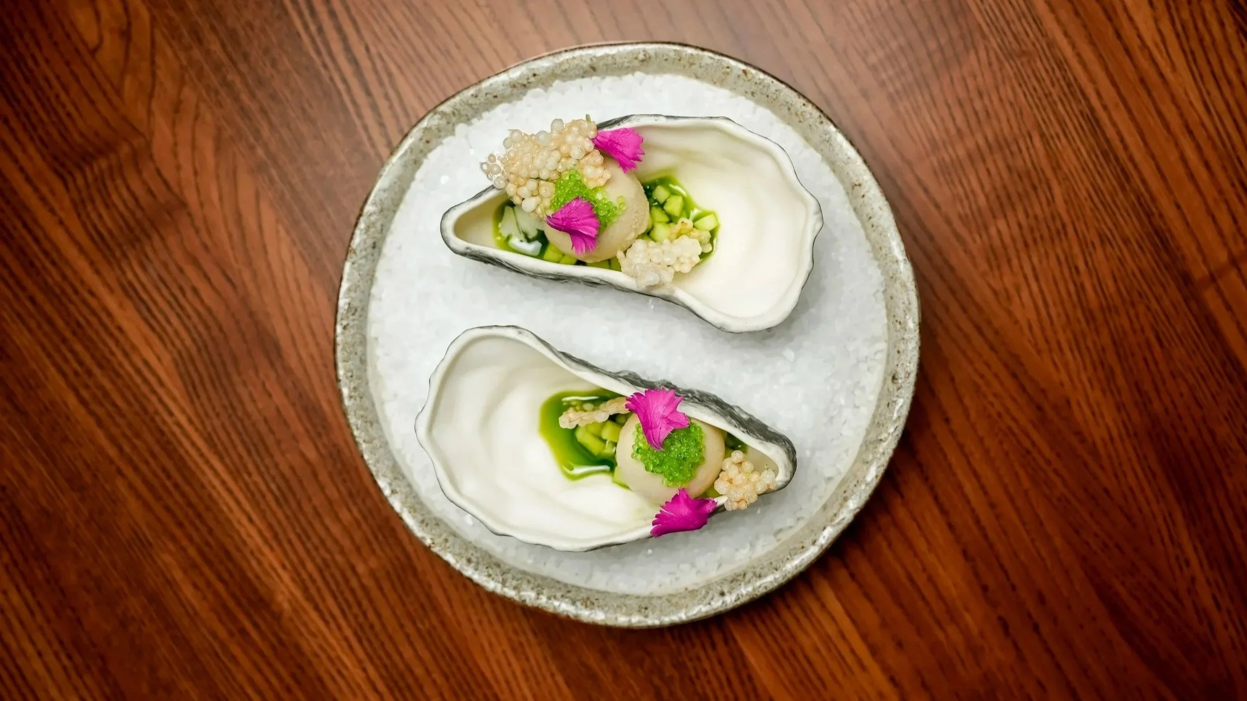 A ceramic plate with two oyster shells filled with a dish garnished with pink flower petals, small cubes, green sauce, and edible beads on a wooden table.
