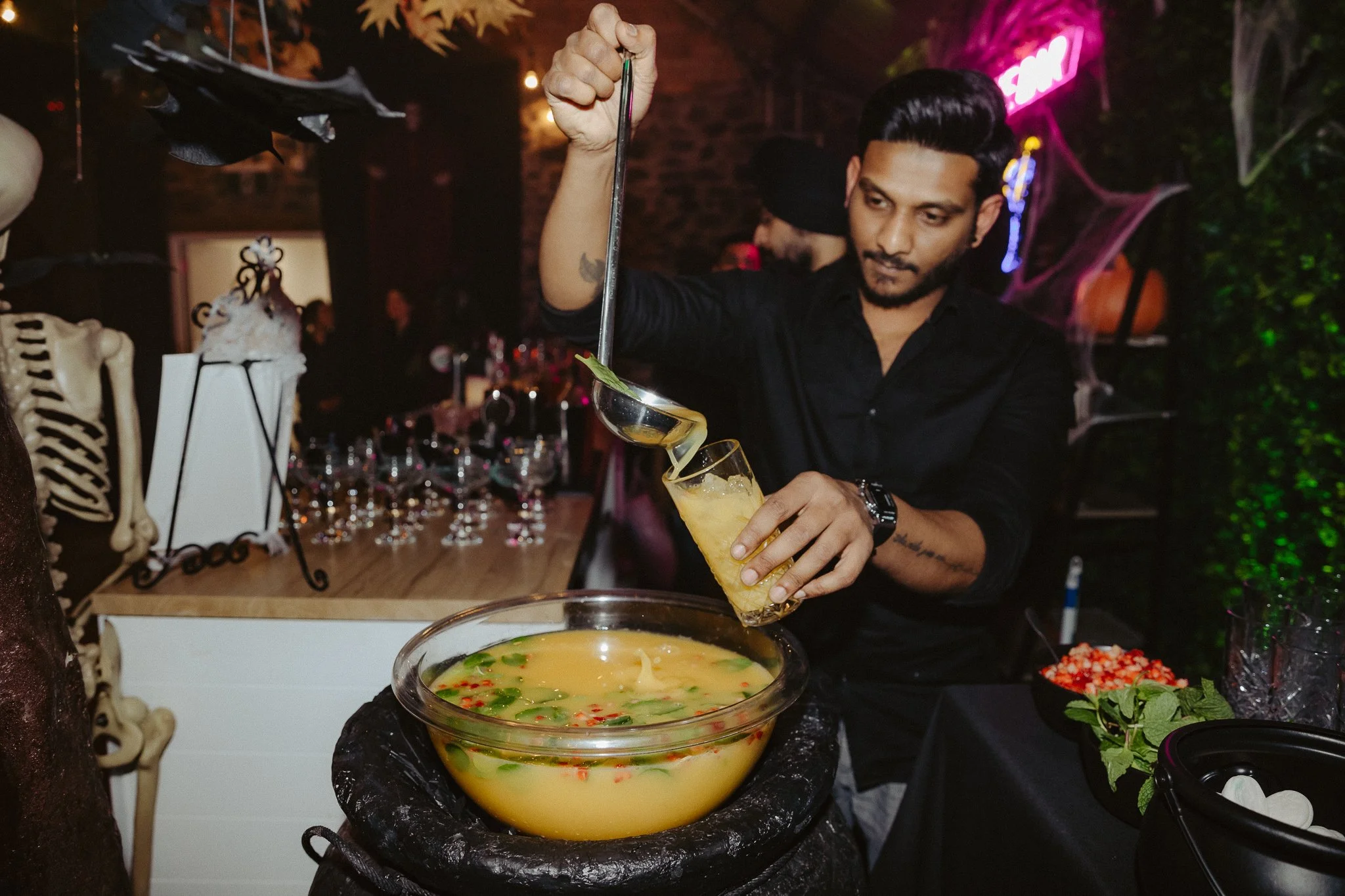 A bartender pours a yellow cocktail into a glass with ice, garnished with a lime wedge, at a Halloween-themed party with skeleton decorations and cobwebs.