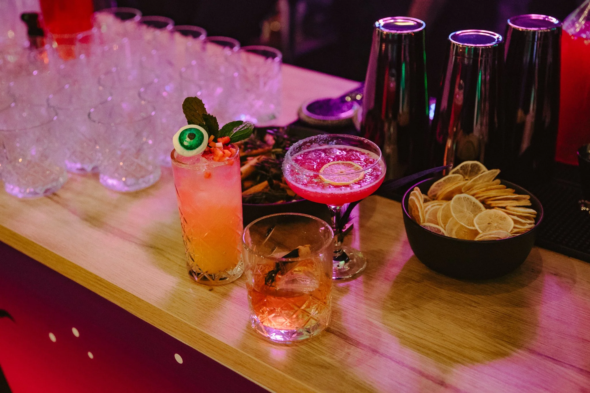 Assorted cocktails on a bar counter with glassware, lemon-lime garnished drinks, lemon slices in a black bowl, and stacked empty glasses.