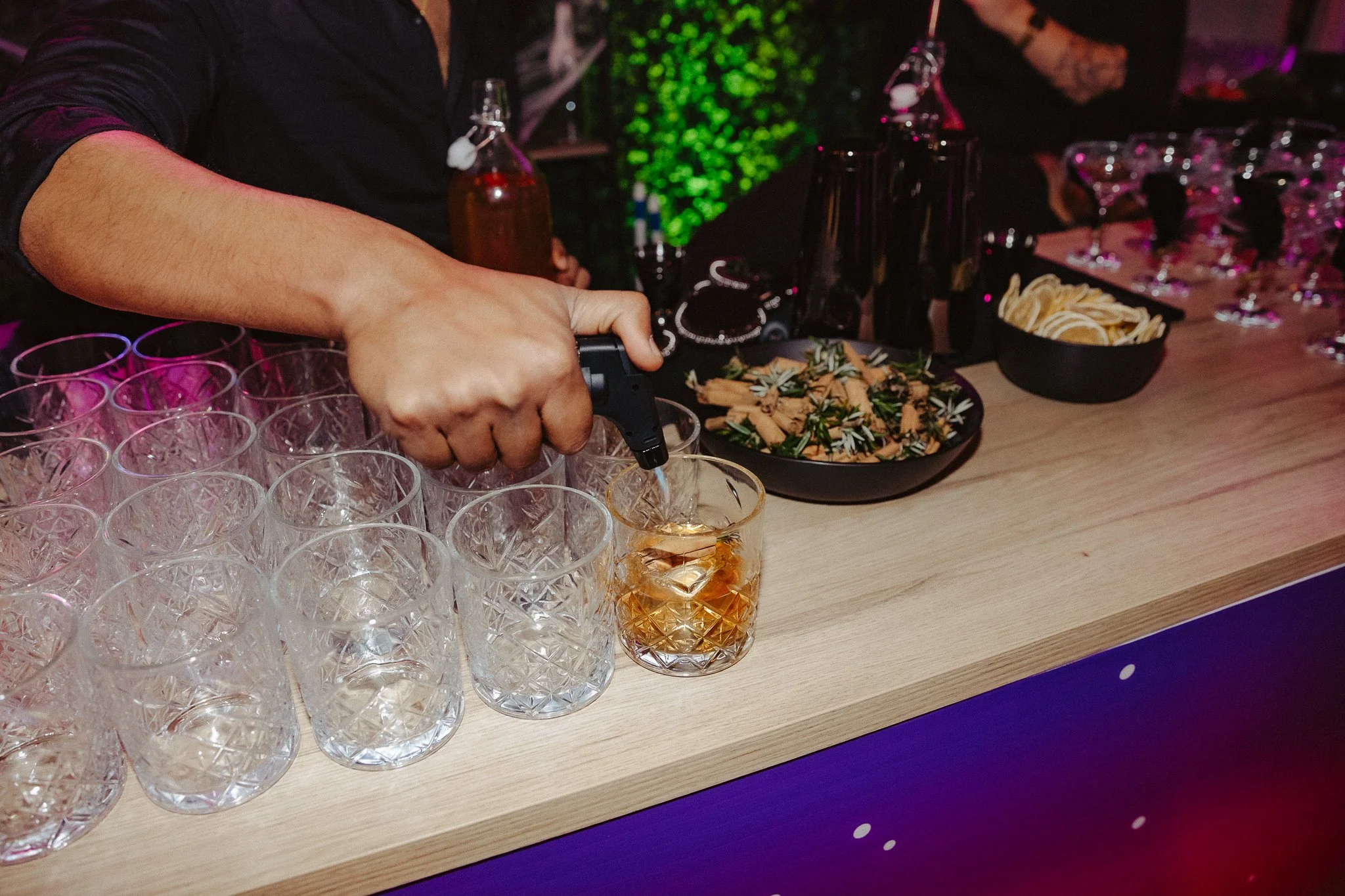 Person pouring a drink into a glass on a bar counter with empty glasses, snacks, and bottles around.